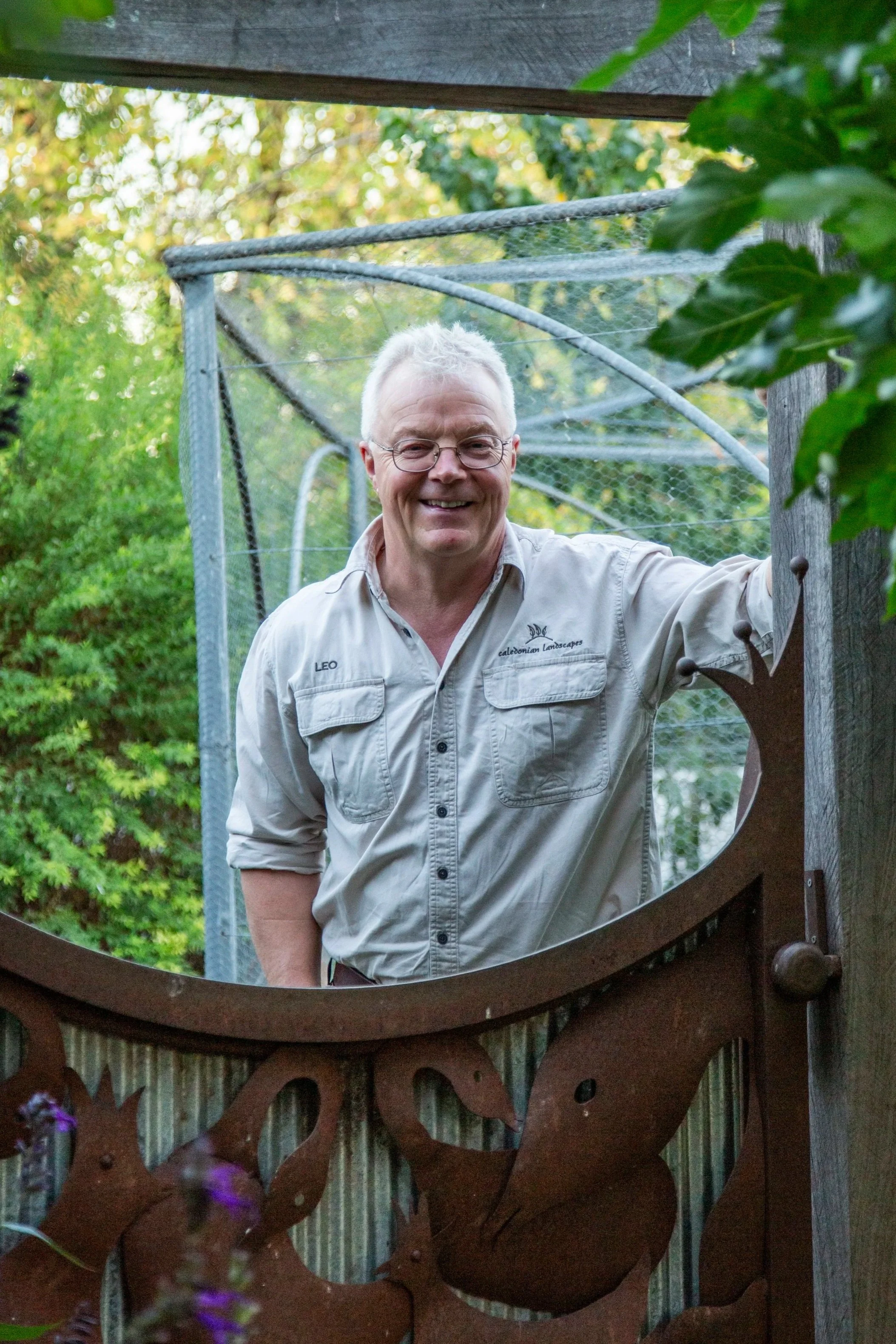 A man with white hair and glasses smiling, wearing a light-colored work shirt, standing outdoors among greenery, seen through a decorative metal garden suncatcher.