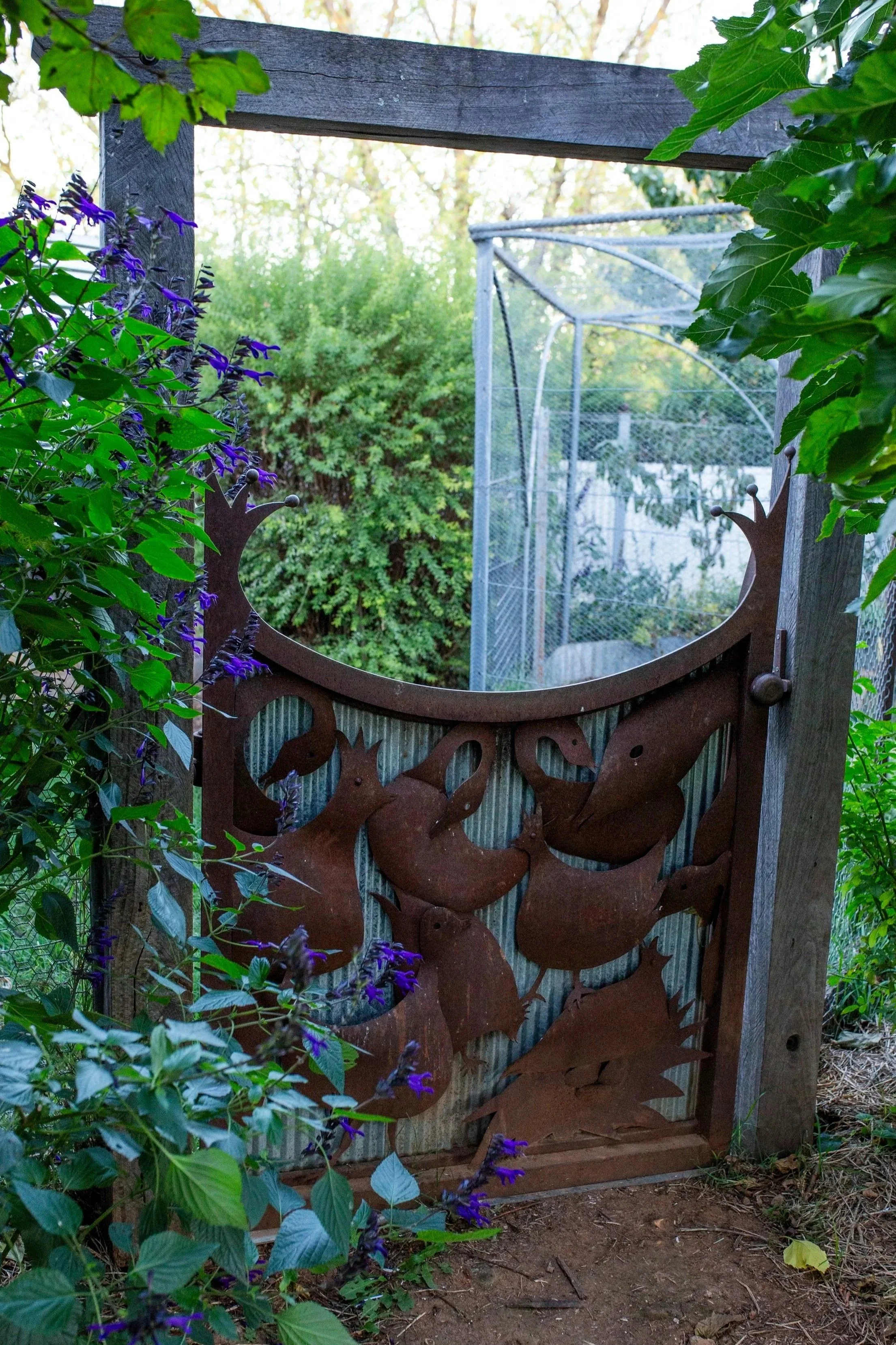 A metal gate with a decorative deer head silhouette design, set on a weathered wooden post fence, surrounded by greenery and purple flowers in a garden setting.
