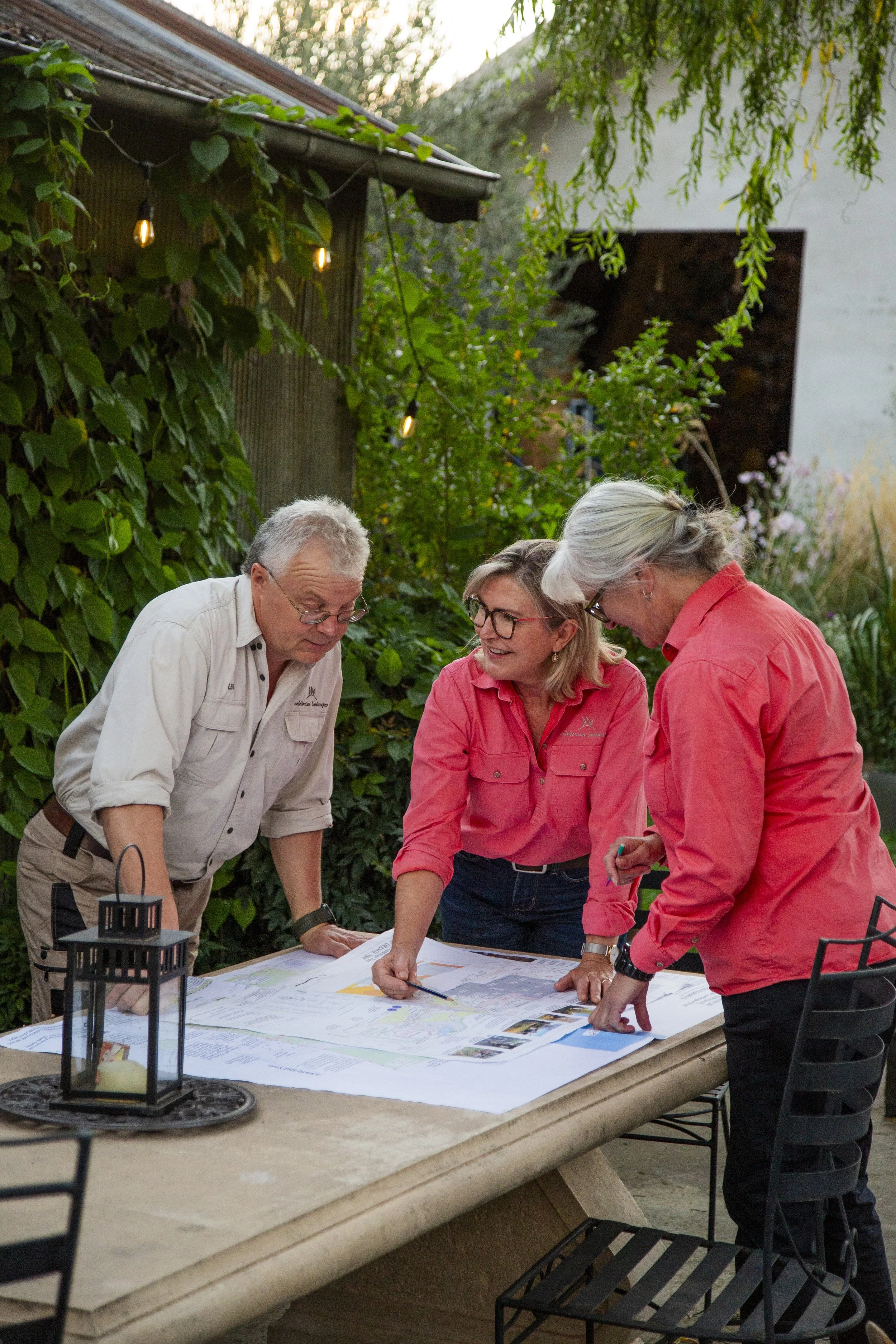 Four people in casual outdoor clothing gathered around a table, examining large maps or plans, with greenery and string lights in the background.