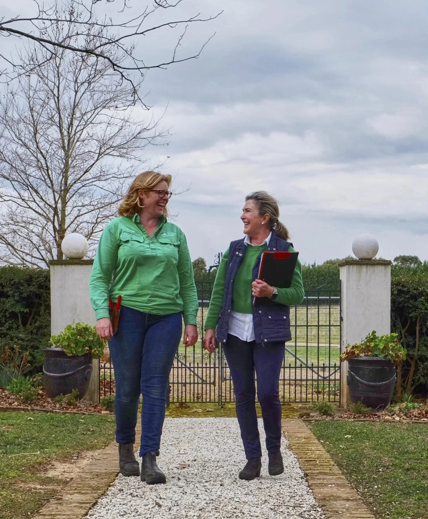Two women walking and talking outdoors near a gate, with potted plants and leafless trees in the background under a cloudy sky.