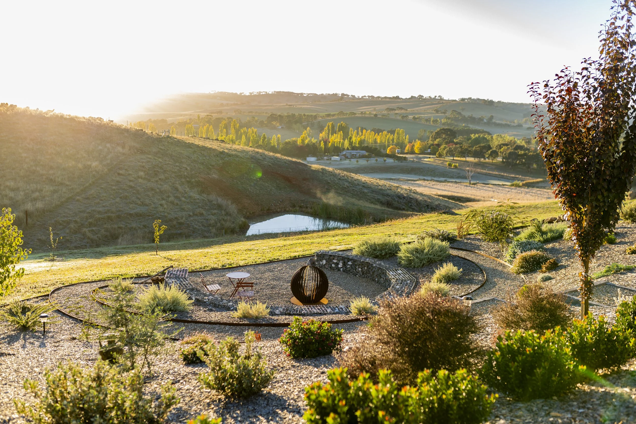 Sunlit rural landscape with rolling hills, trees, small pond, and outdoor seating area with chairs and a spherical fire pit.
