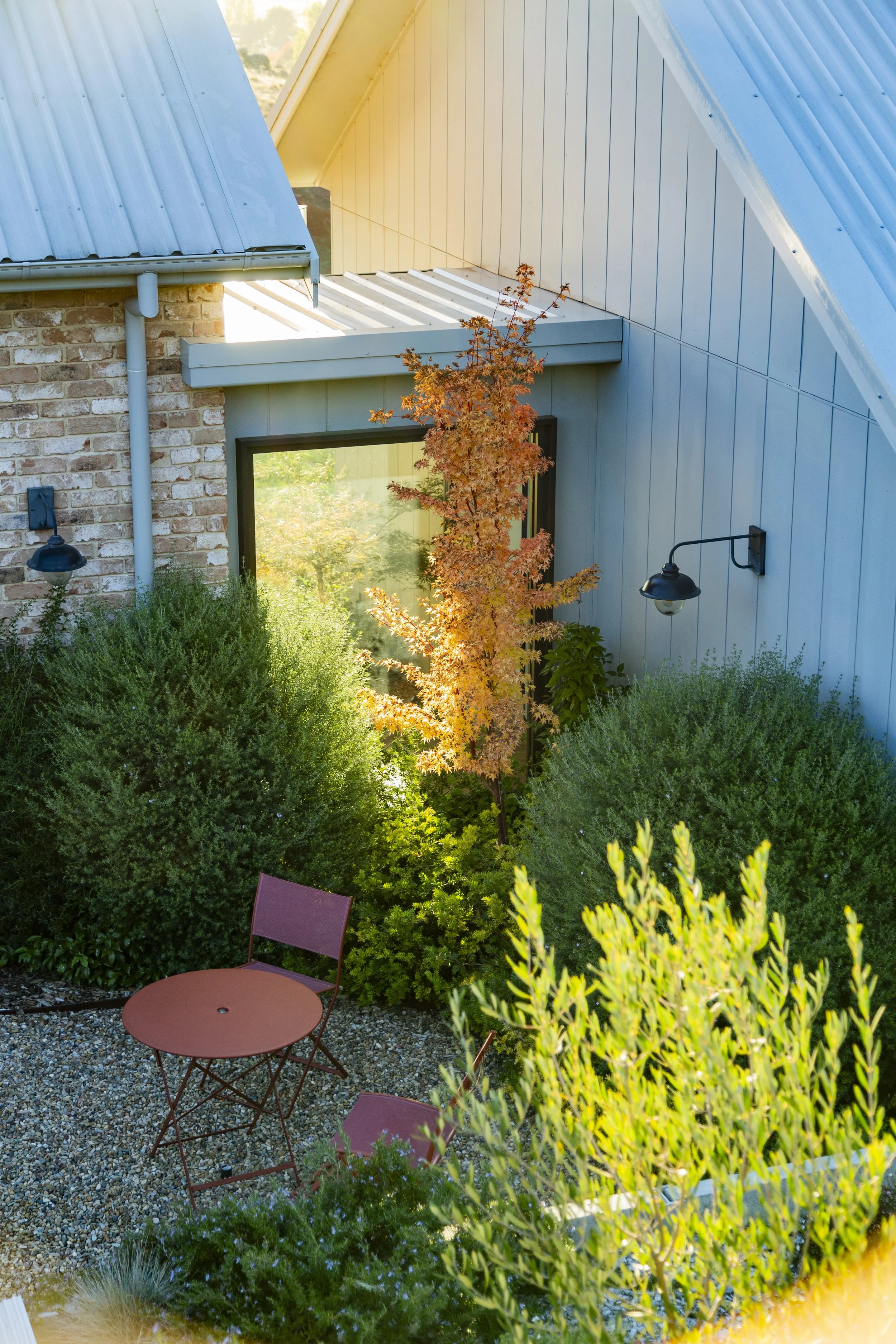 A backyard garden with a small round metal table and two matching chairs on a gravel surface, surrounded by lush green bushes, trees, and a modern house with a sloped metal roof and large windows.