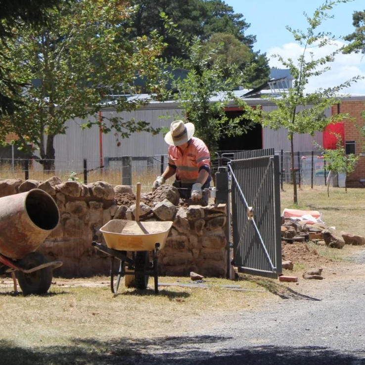 Worker building a stone wall with rocks, using a wheelbarrow and tools, outdoors in a sunny area.