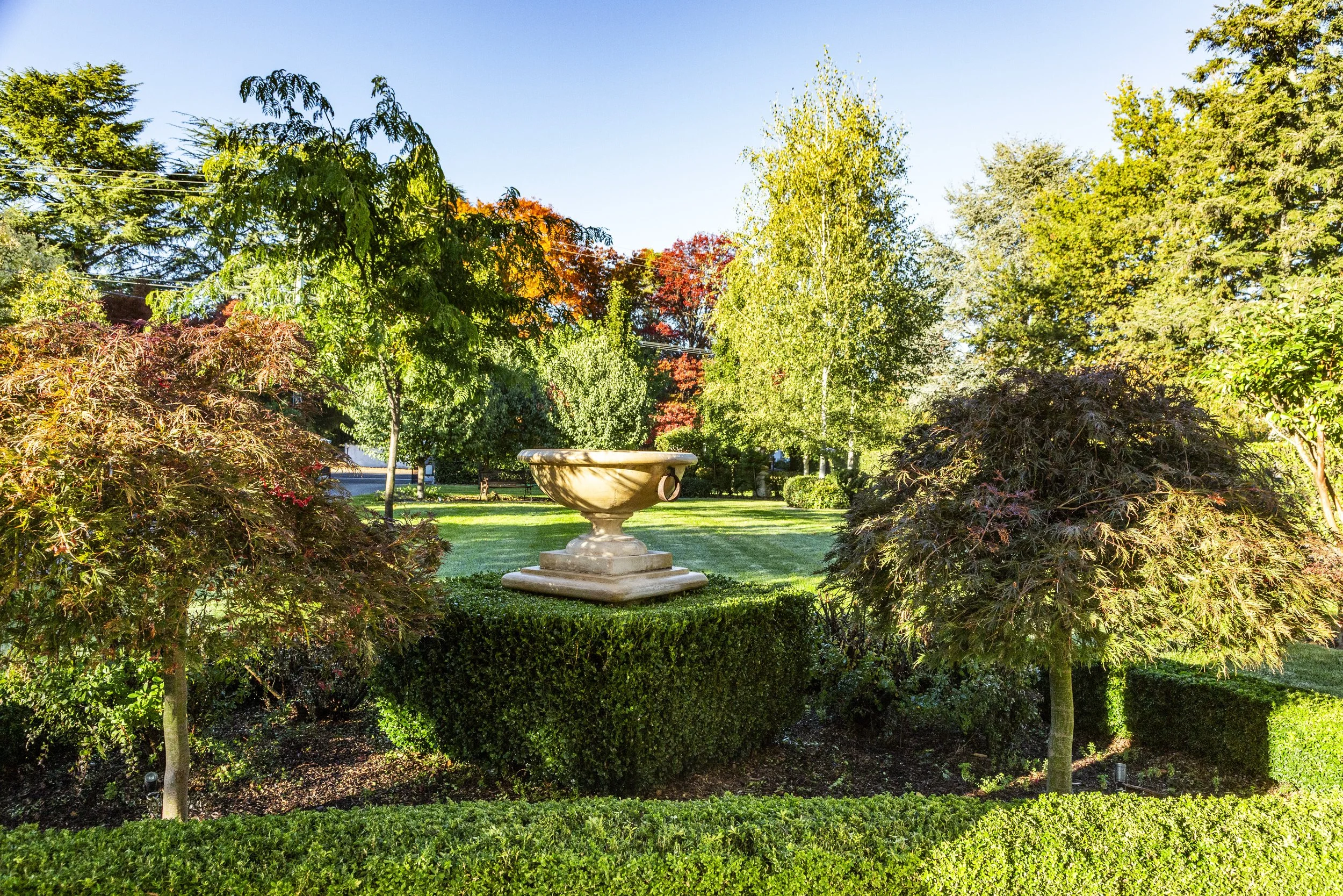A landscaped park with a variety of trees, a stone fountain in the center, and well-maintained grass and bushes, under a clear blue sky.