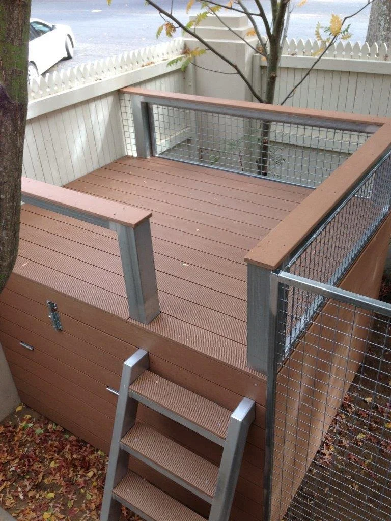 Small outdoor deck with wood planks, metal railing with wire mesh, a step ladder, and a white picket fence in the background with trees and a parked white vehicle.