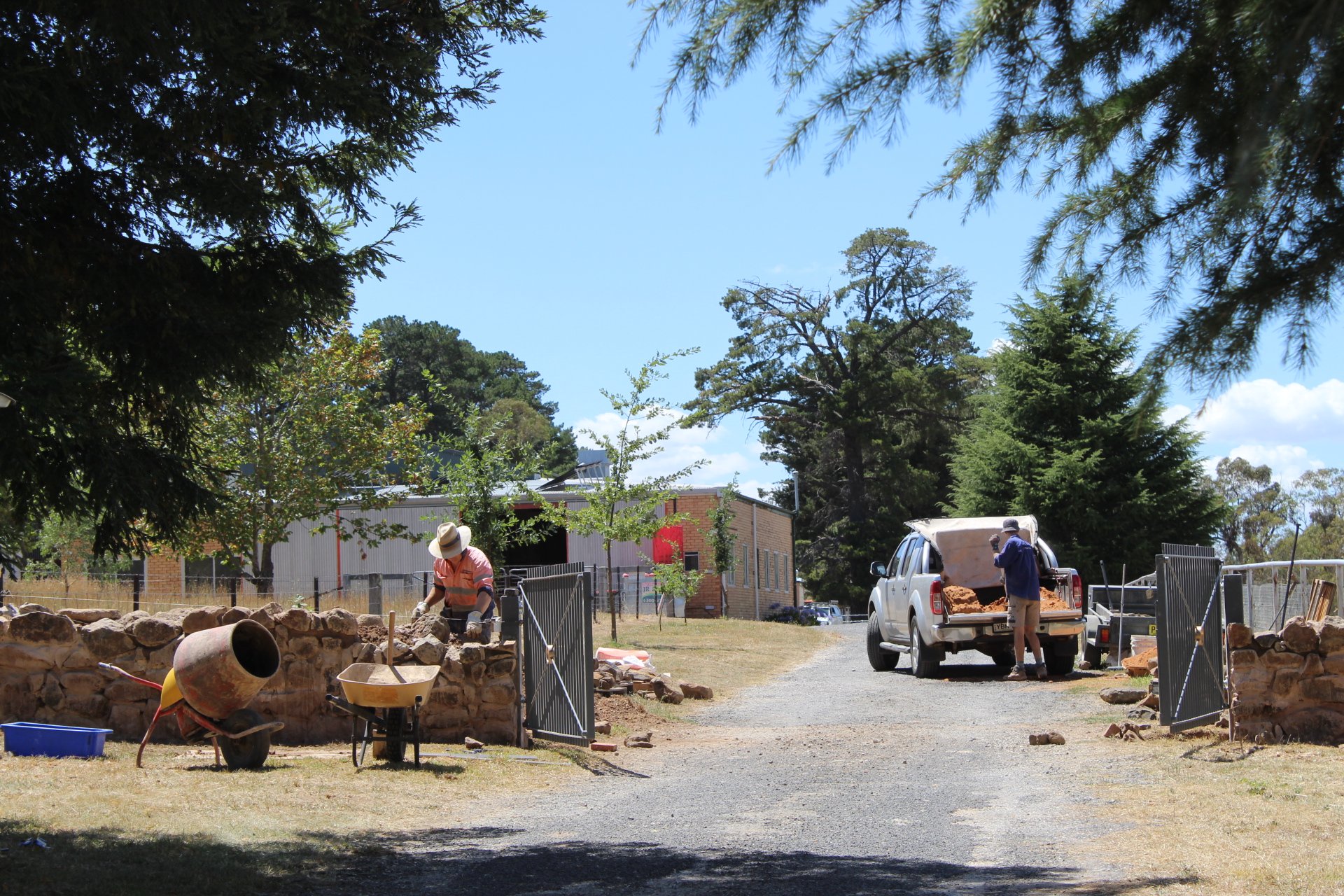 People working outdoors on a construction project with trees and buildings in the background, a white pickup truck with construction materials, and various tools and equipment on the ground.
