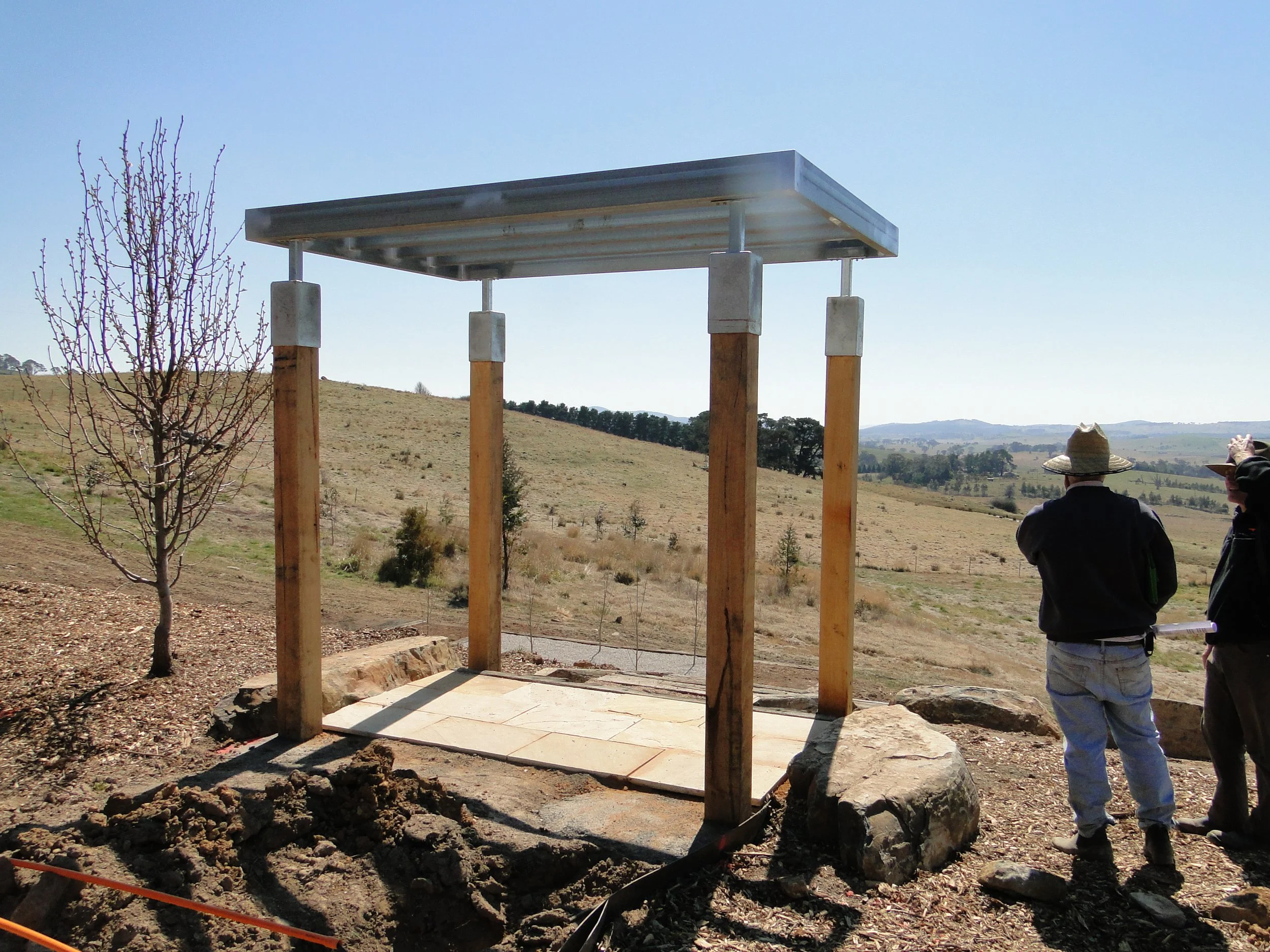Construction site with a metal roof structure supported by four wooden posts, set in a rural landscape with rolling hills and some trees, two people wearing hats and jackets walking nearby.