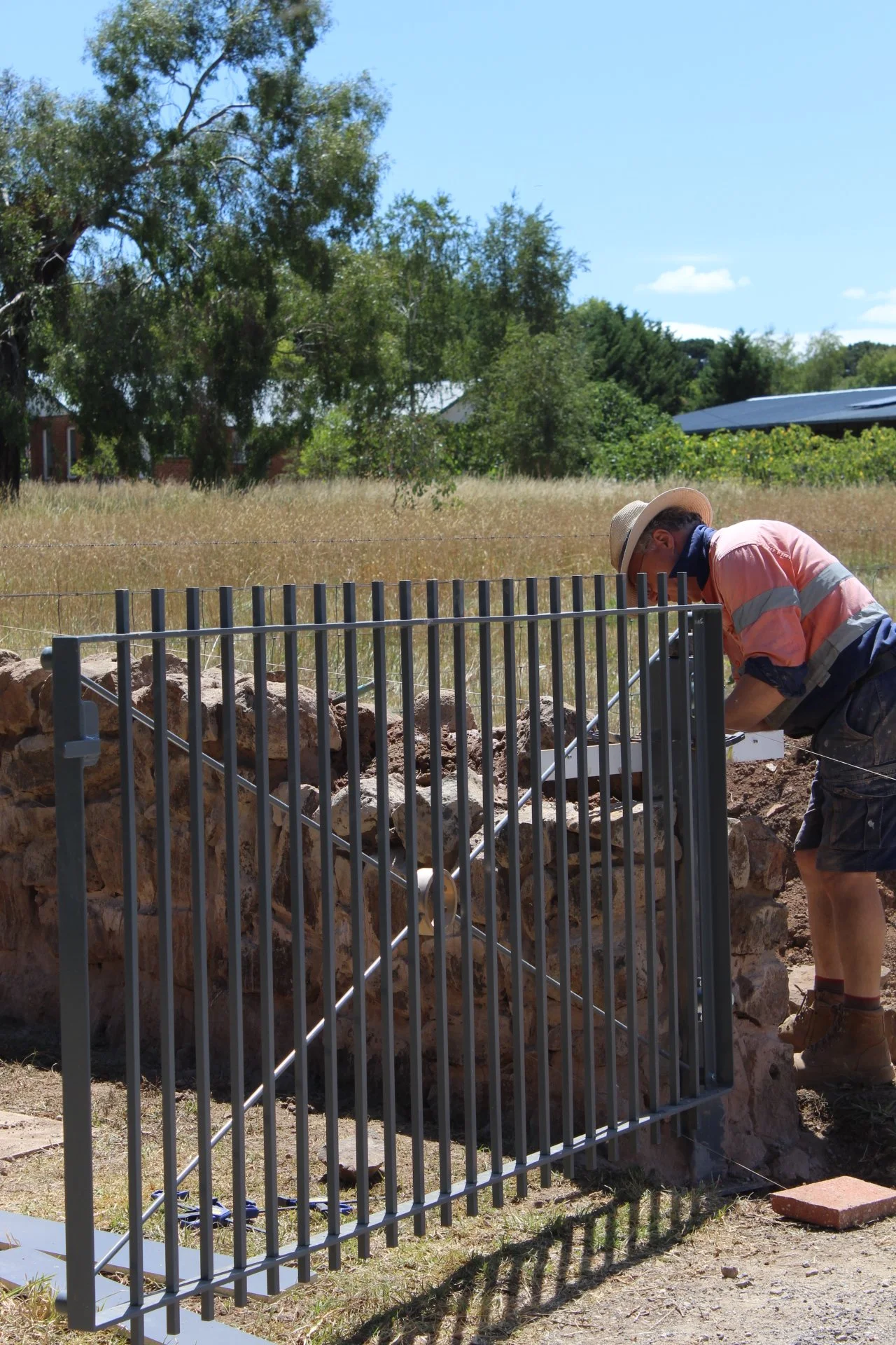A person wearing a hat, sunglasses, a face mask, a pink shirt, and shorts installing a metal gate on a stone wall in an outdoor area with trees, grass, and a blue sky.