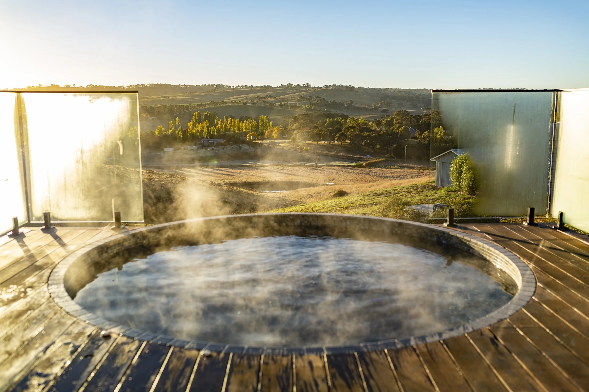 Hot tub with steam rising, overlooking a scenic countryside landscape with rolling hills, trees, and a clear sky at sunrise or sunset.