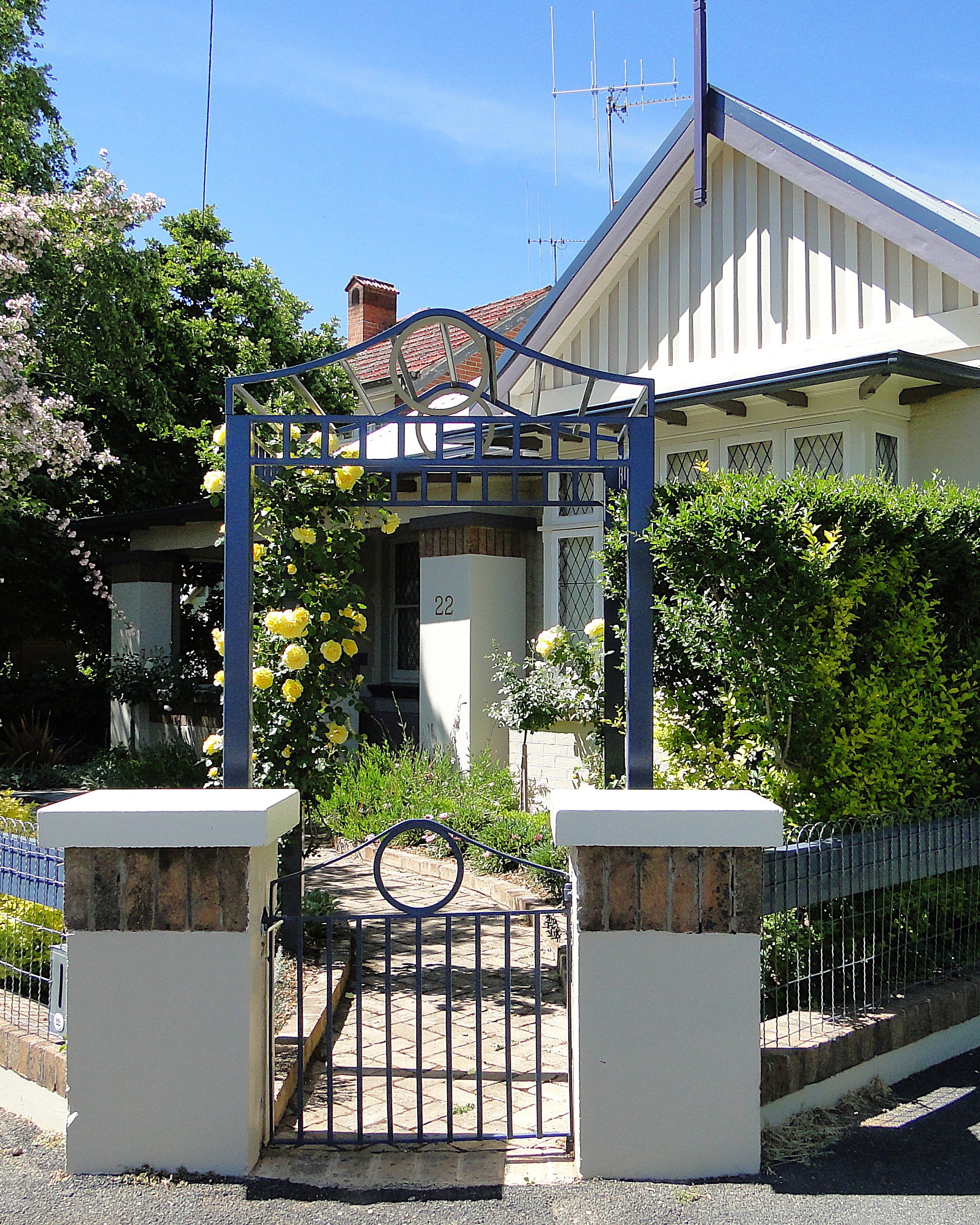 Front view of a house with a small gate, pillars, lush greenery, pink and yellow flowers, and a pathway leading to the entrance.
