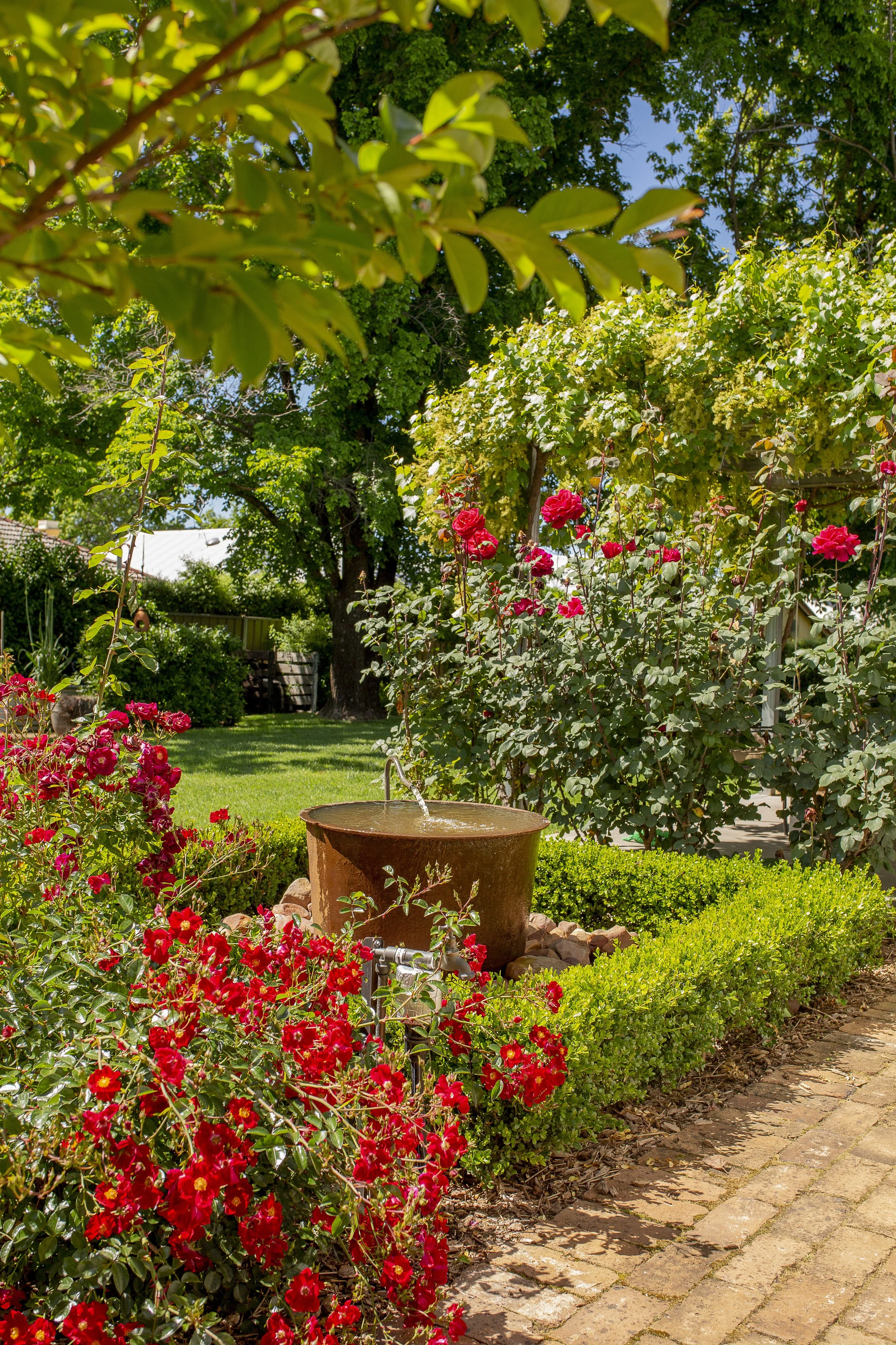 A vibrant garden scene with red flowers, green bushes, and trees, featuring a rusty water fountain and brick pathway under a bright blue sky.