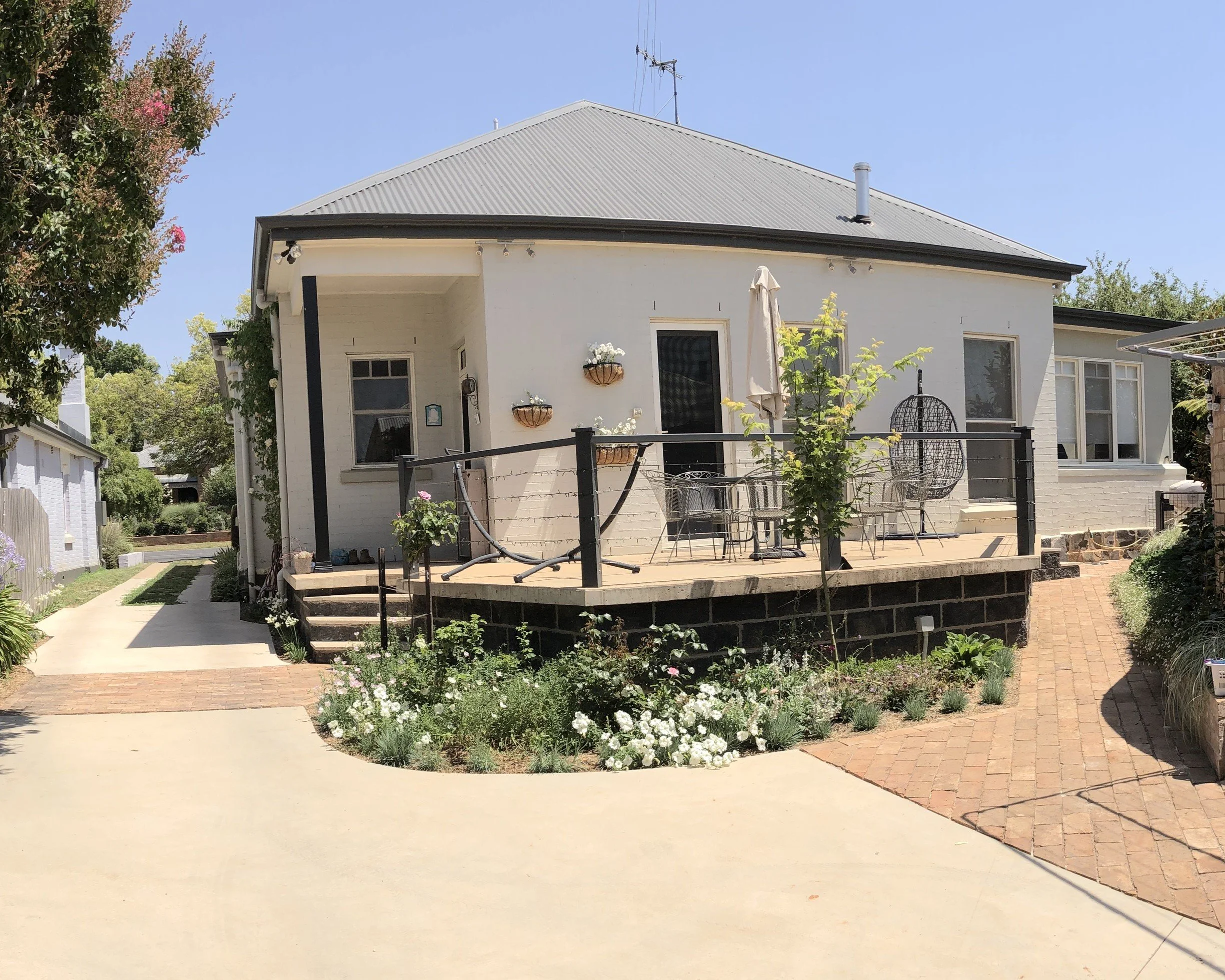 A white house with a gray metal roof, a deck with outdoor furniture, potted plants, and a garden with flowers and trees, under a clear blue sky.