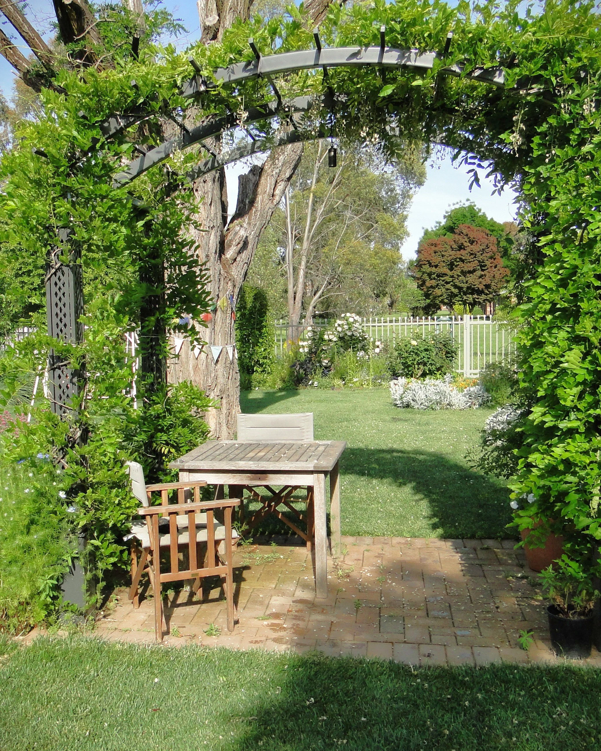 A backyard garden scene featuring a wooden outdoor table and chairs, a green hedge archway, trees, a white picket fence, and various plants and flowers under a blue sky with scattered clouds.