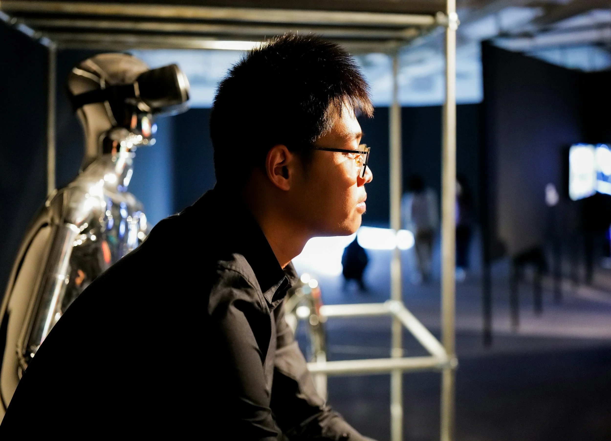 A person with short dark hair and glasses sitting in profile facing right, with a robot-like figure and a dark background with some light sources.