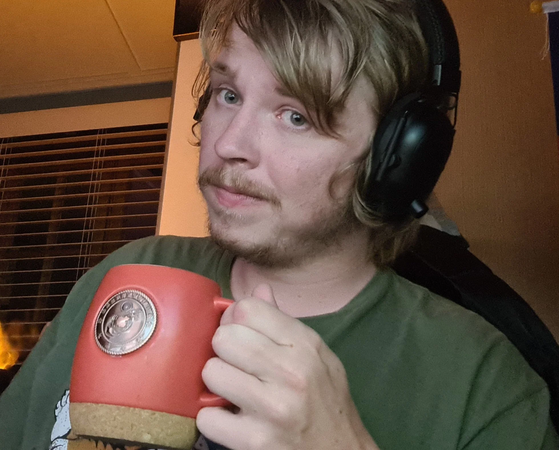 A young man with long blonde hair, a mustache, and a beard wears large black over-ear headphones and holds a red boxing glove in front of him, looking at the camera. The background features wooden window blinds and a beige wall.
