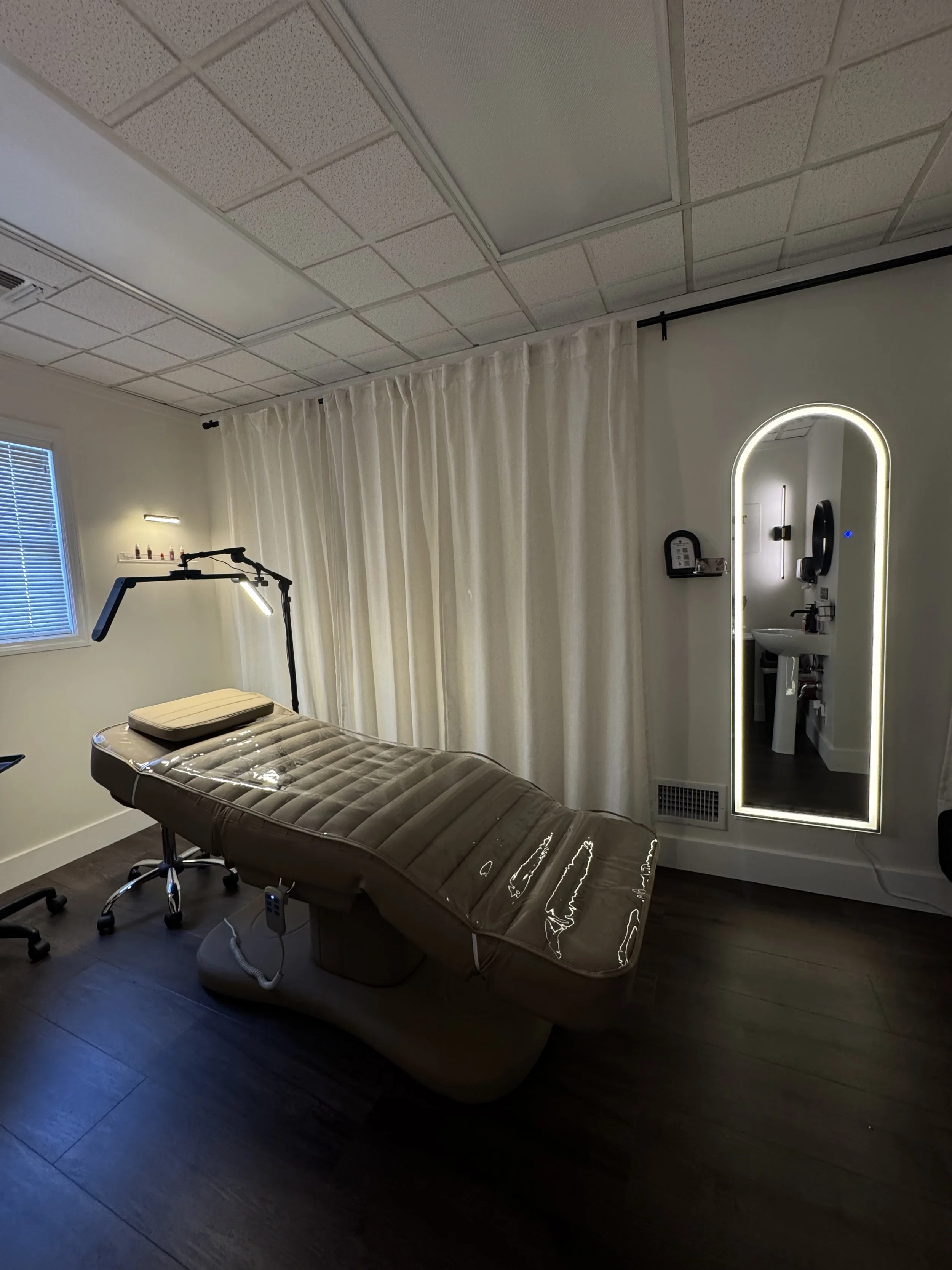 A clinical room with a medical examination table, a standing magnifying lamp, a window with blinds, and a large mirror with LED lighting around the frame. There are also some small bottles on a shelf near the window and a curtain dividing the room.