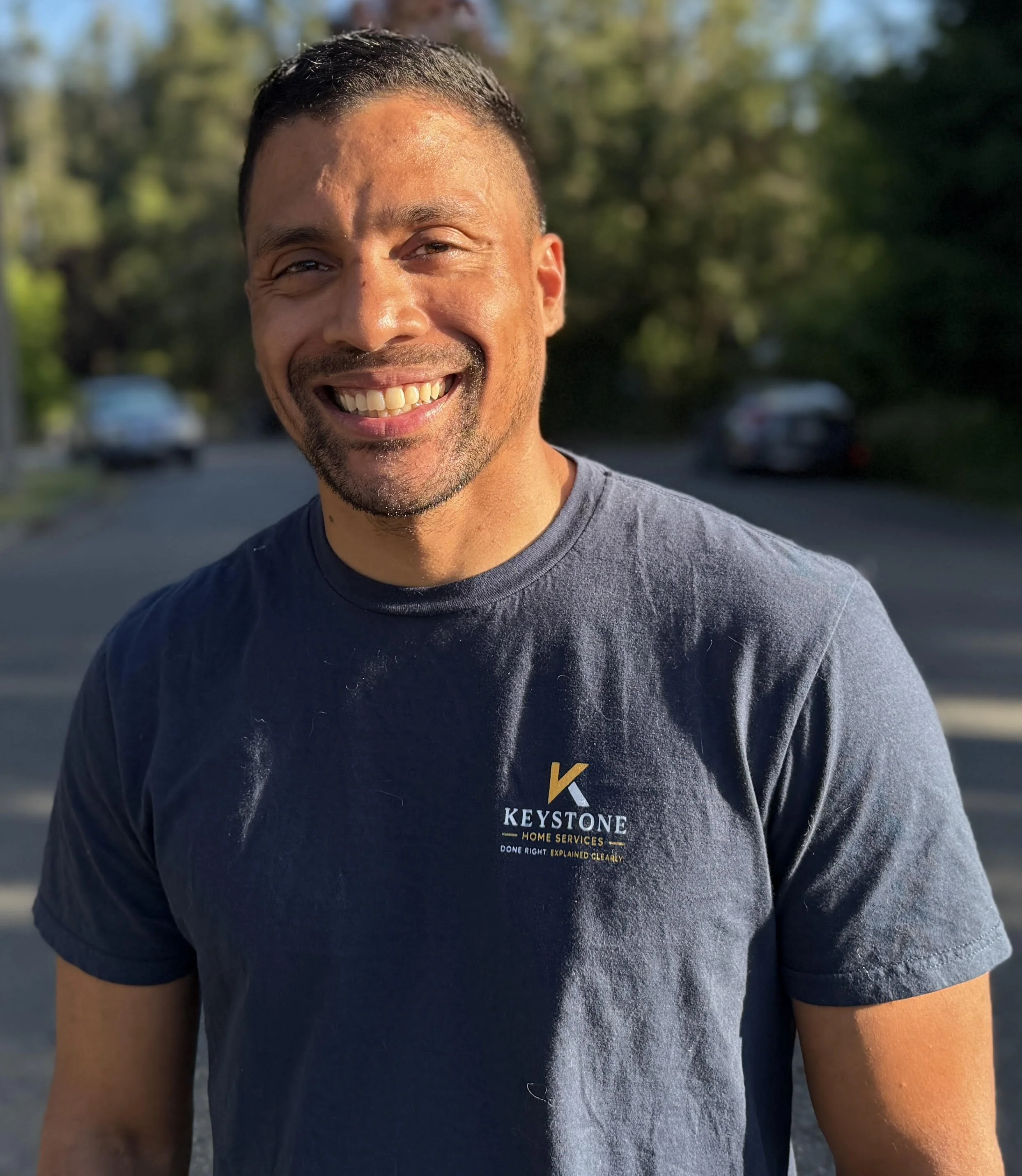 Close-up of a smiling man wearing a black Keystone Home Services t-shirt, standing outdoors with blurred trees and cars in the background, during daylight.