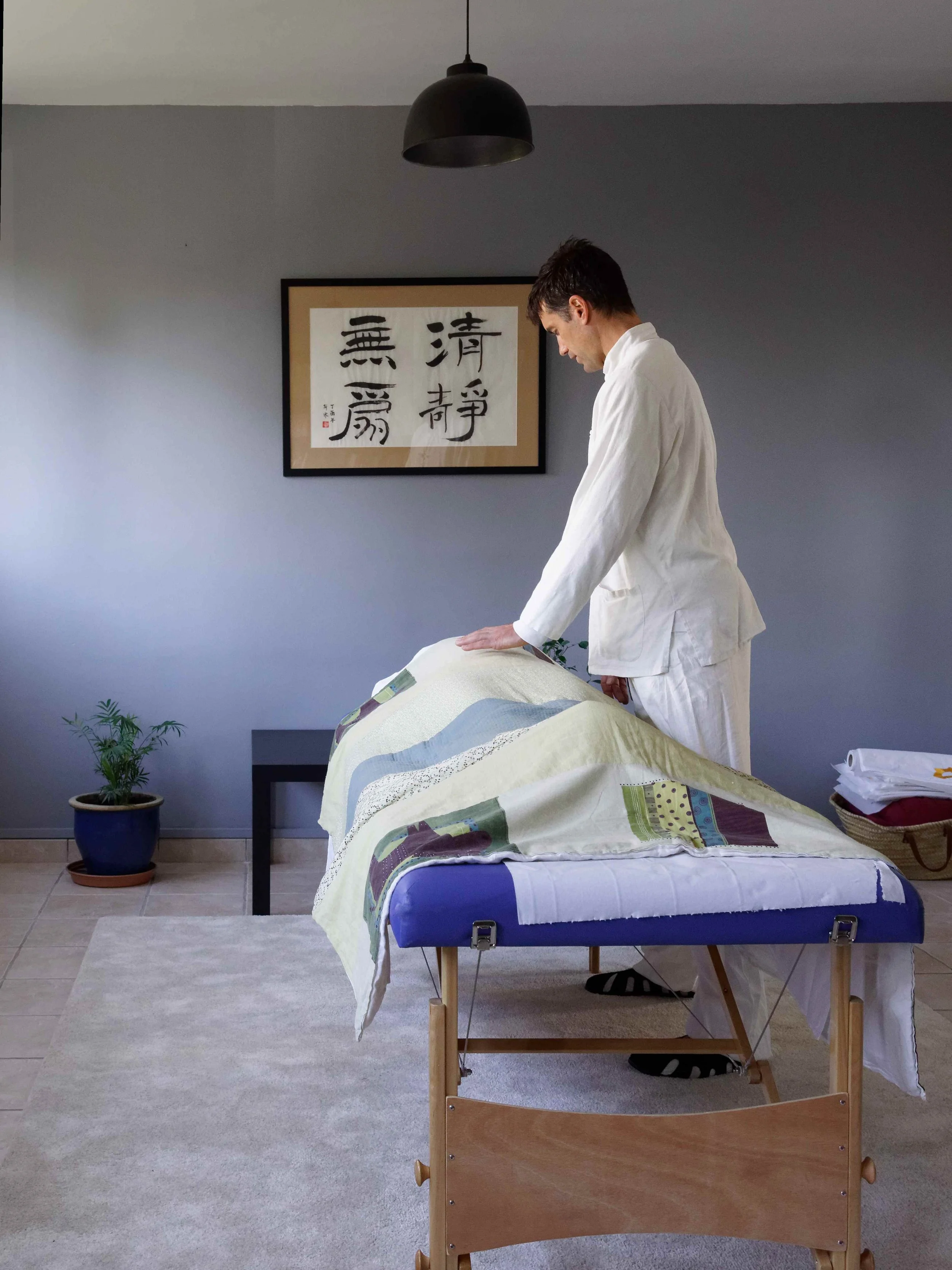 Une pièce avec un lit de massage, une personne en tenue blanche effectuant un massage, une peinture avec des caractères chinois sur le mur, une plante en pot, et une table avec des papiers. La pièce a un décor simple et moderne.