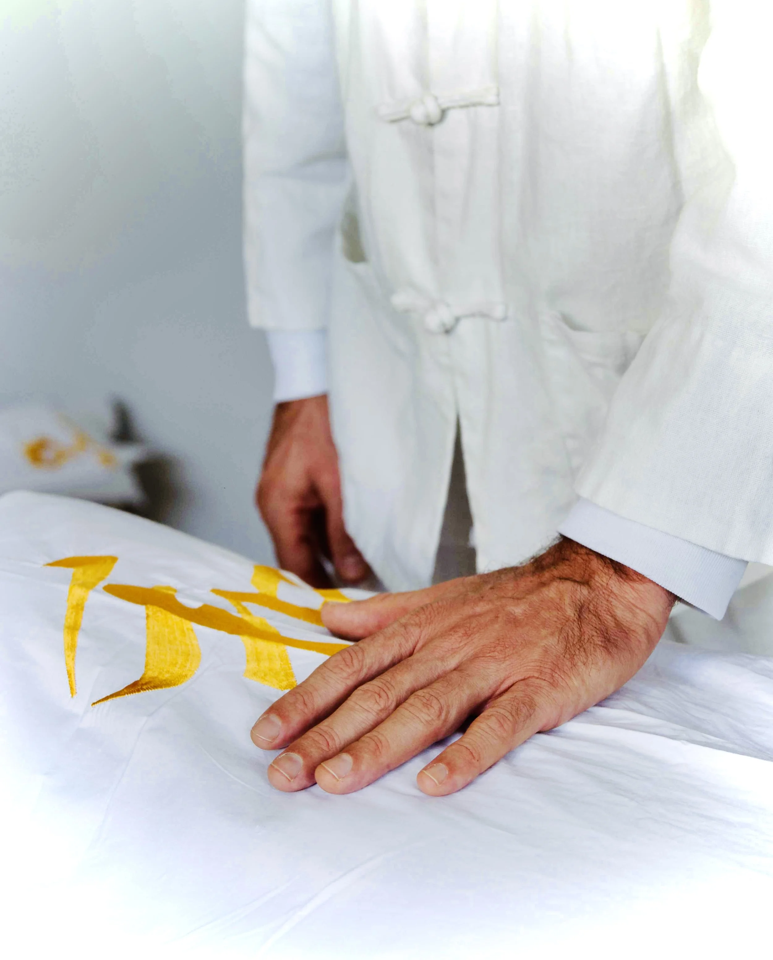 Une personne en vêtements blanc avec une manche retroussée pose sa main sur un lit ou une surface blanche, avec un drap ou un tissu blanc avec des lettres jaunes dessus.