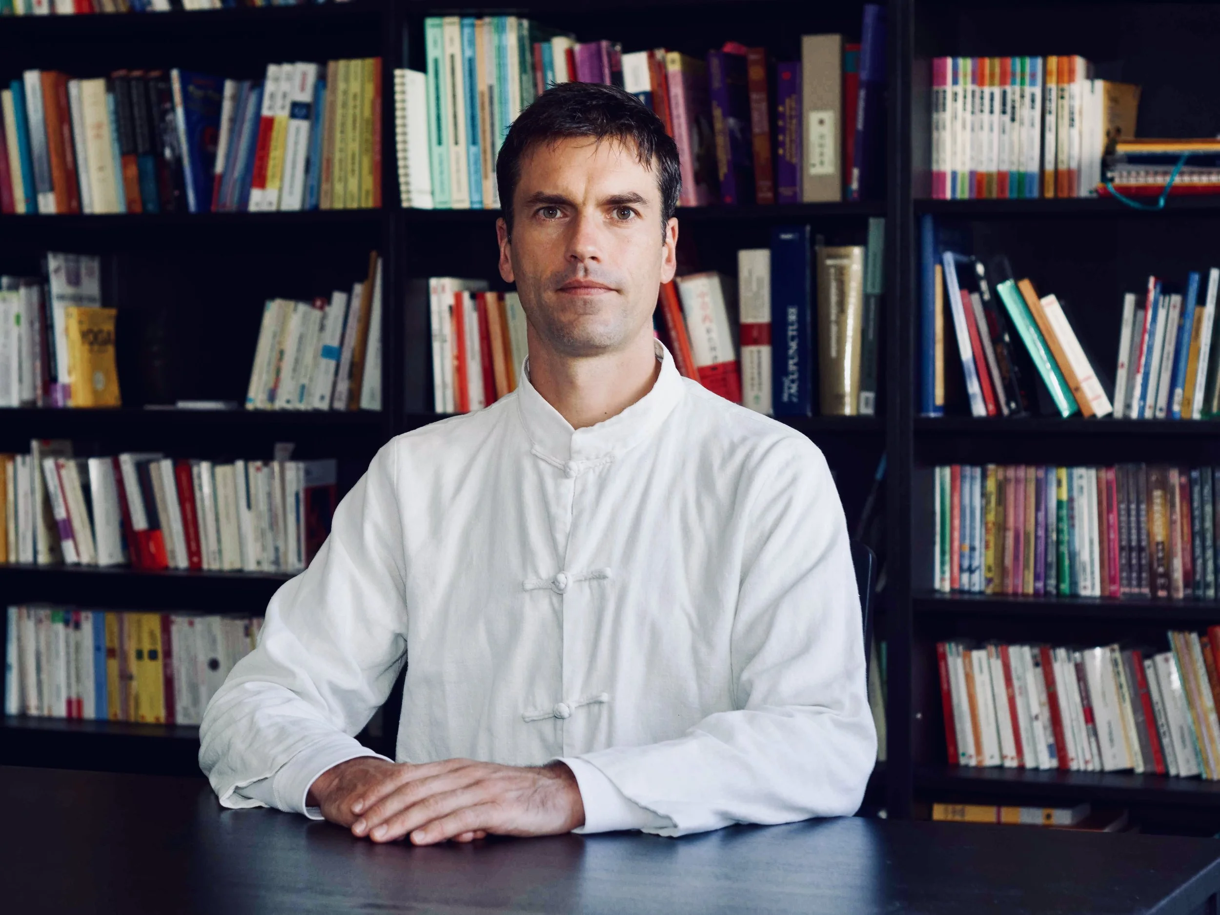 Homme en uniforme de cuisine, assis à une table devant une bibliothèque pleine de livres.