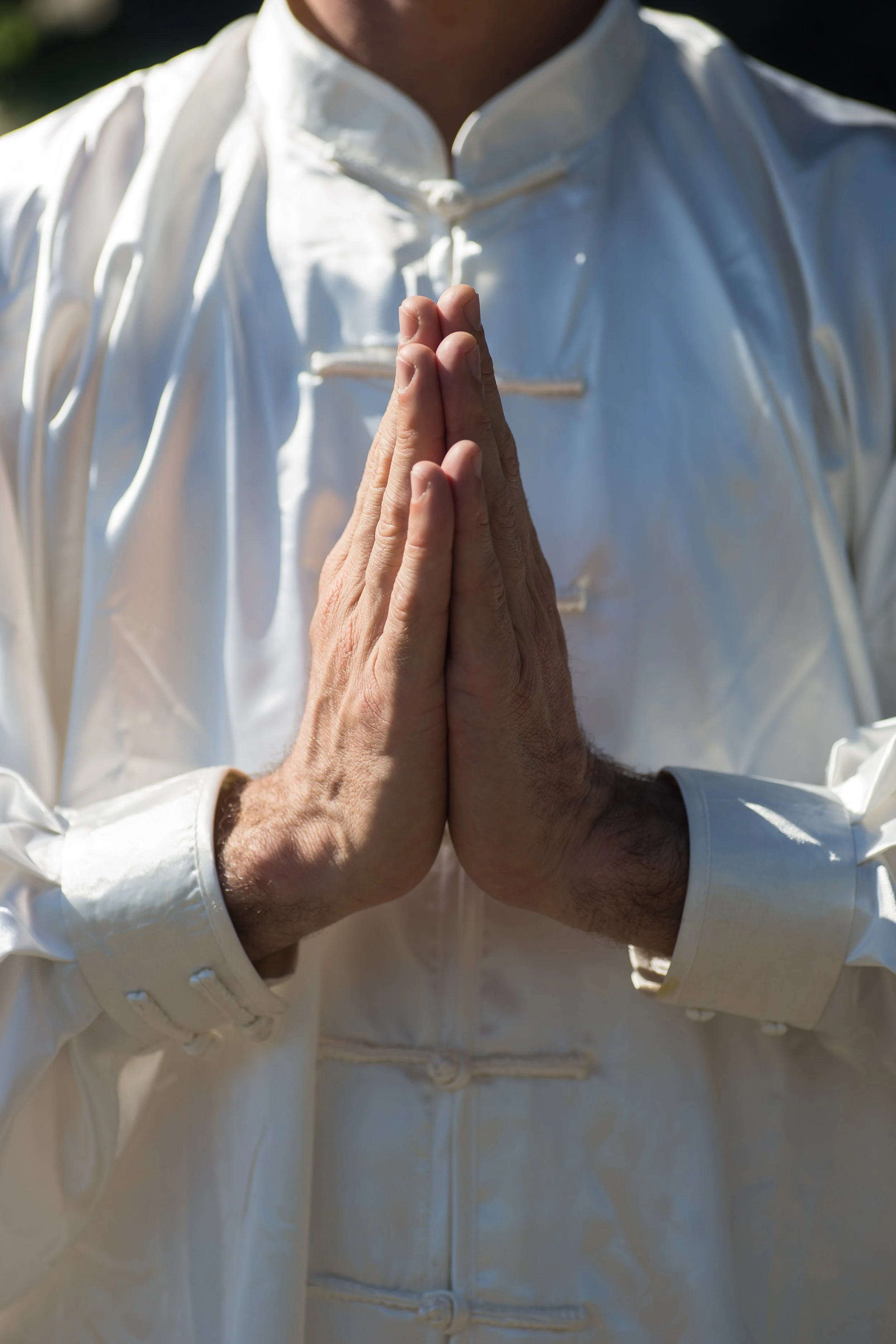 Une personne porte un kimono blanc avec ses mains jointes en position de prière ou de méditation. La photo est prise en extérieur, face au soleil.