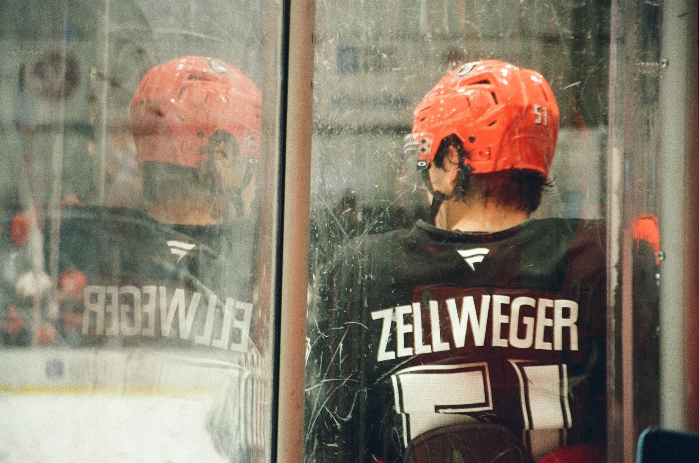 A person wearing a red hockey helmet and a black shirt with white text, standing behind a glass window with visible scratches and smudges, with the reflection of the helmet in the glass. The setting appears to be an ice rink or hockey arena, and the 