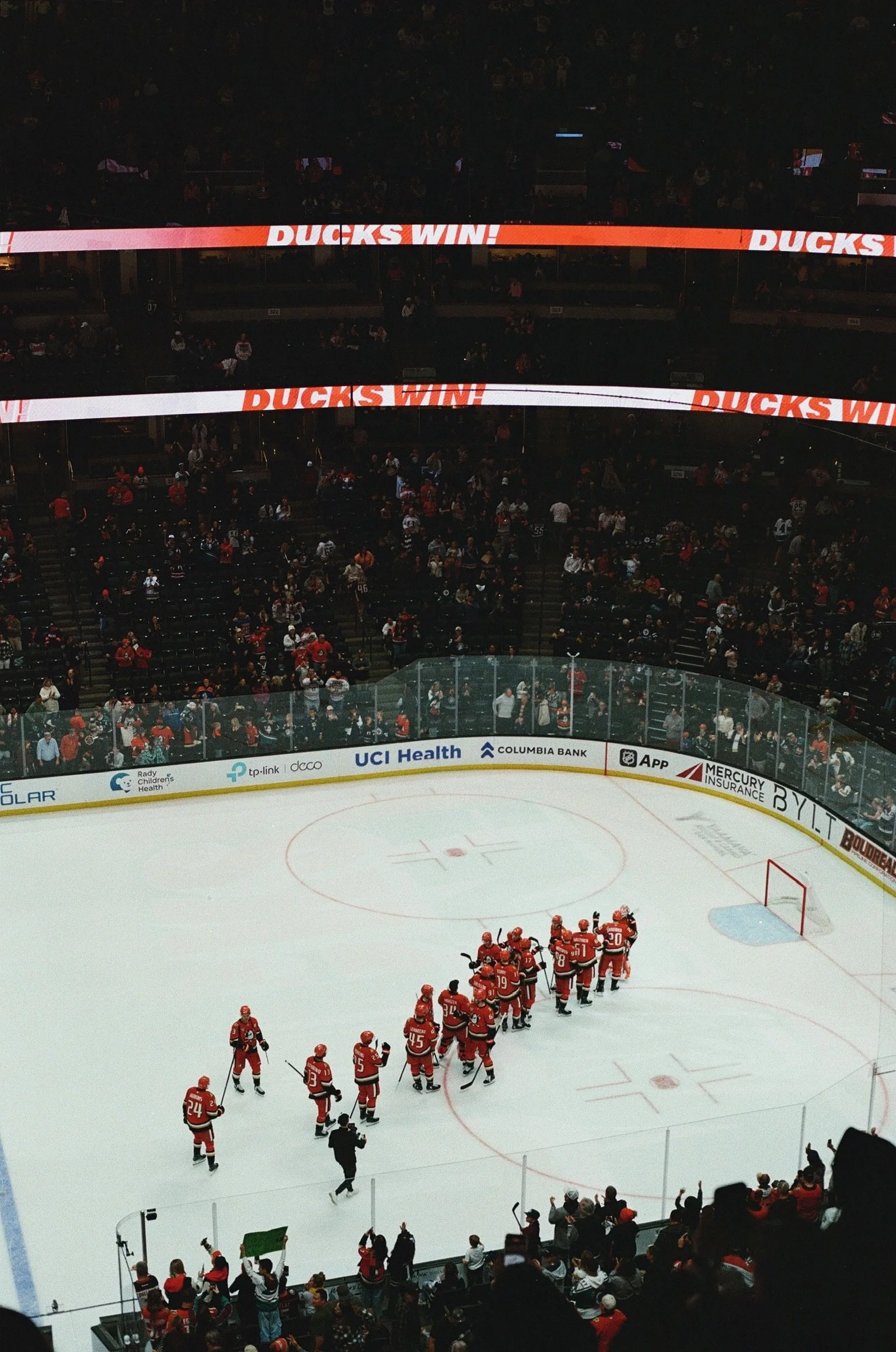 Ice hockey players in red jerseys on the ice rink with spectators in the stands and a large digital banner displaying 'DUCKS WIN!'