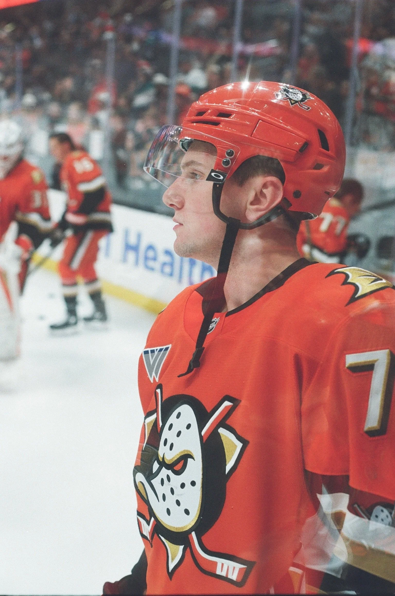 Close-up of a hockey player in an orange Anaheim Ducks jersey and helmet, with other players and spectators in the background.