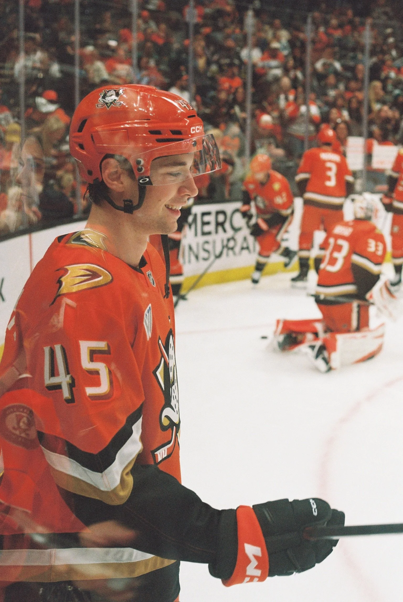 Hockey player in orange jersey smiling on the ice, crowd in stands and other players in background.