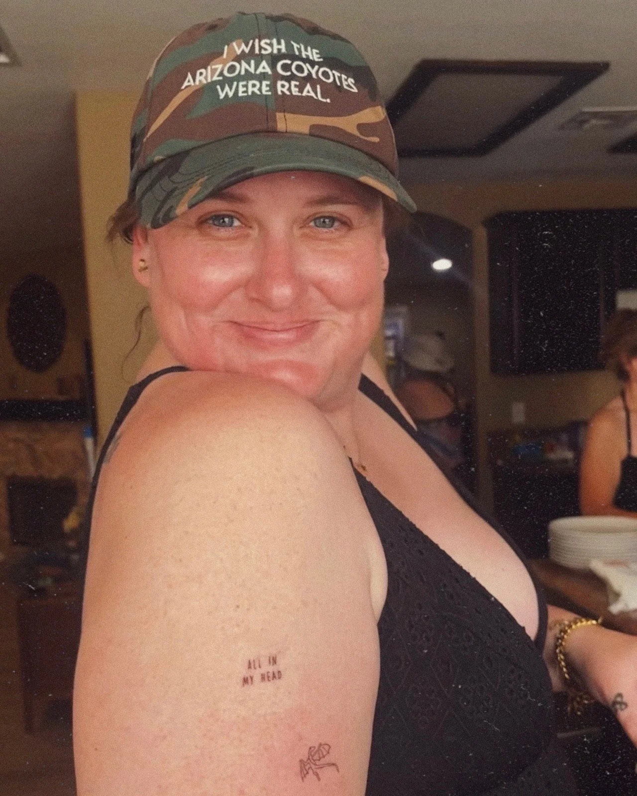 A woman wearing a camouflage cap with the text "I wish the Arizona coyotes were real". She is smiling and has tattoos on her arm, including one that says "All in my head". She is wearing a black tank top and is indoors.