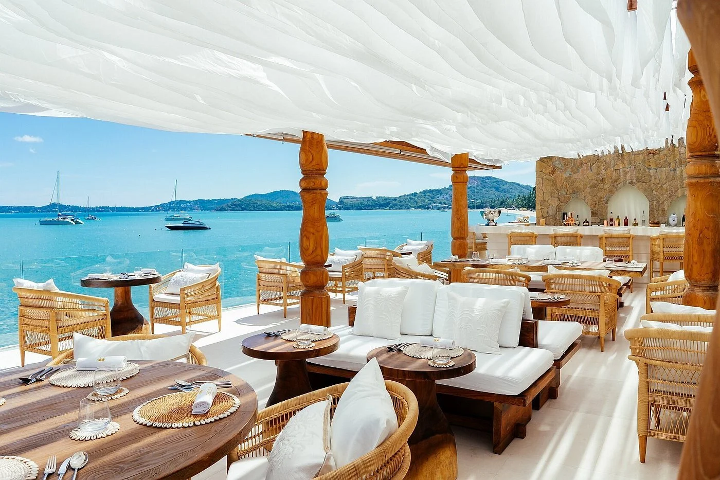 Upscale seaside restaurant with wooden furniture, white cushions, and a view of the ocean with sailboats and a hilly landscape in the background.