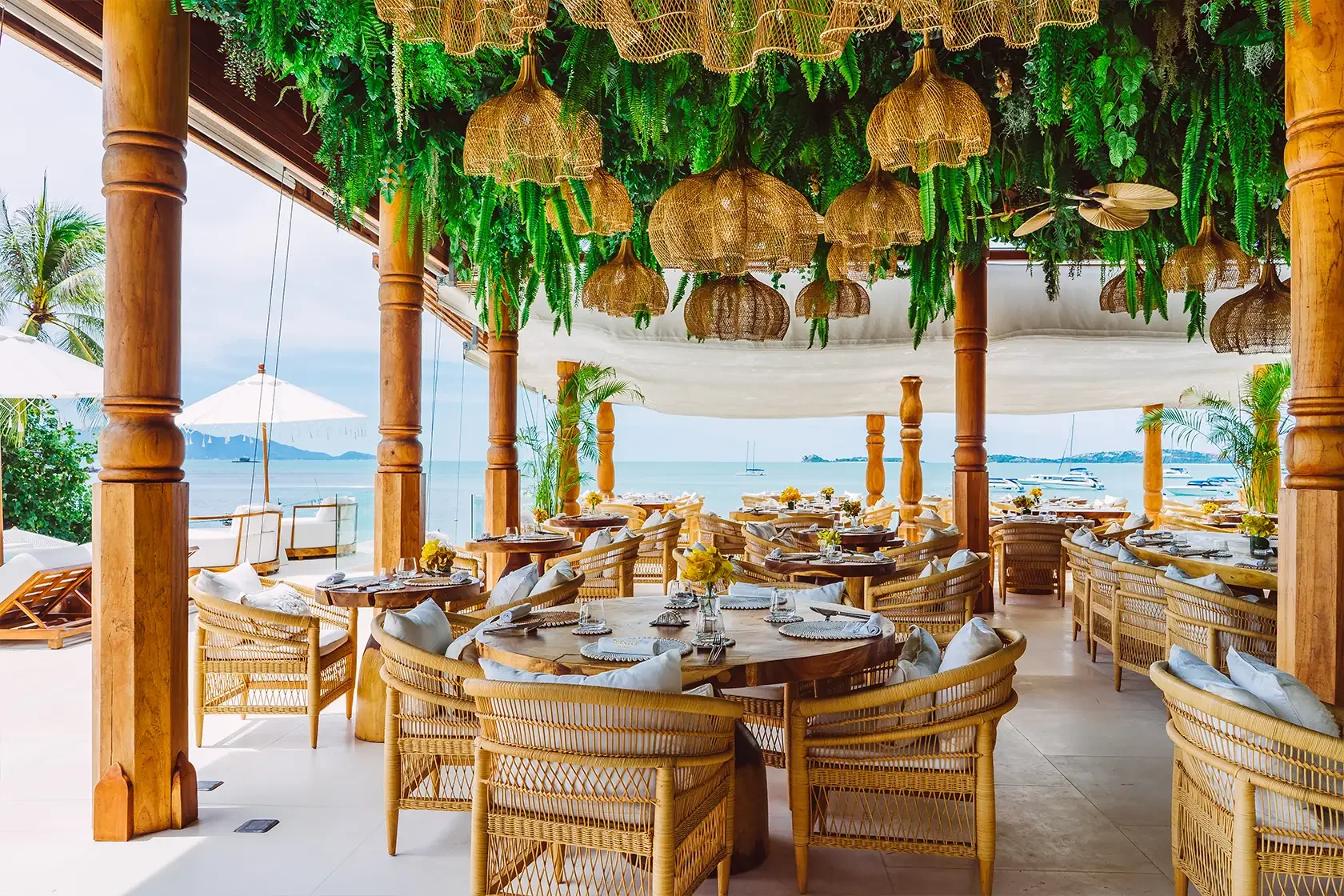 Open-air seaside restaurant with round wooden tables and rattan chairs, decorated with white cloth napkins and yellow flowers. The ceiling has hanging wicker lamps and lush green foliage. In the background, there is a view of the ocean with sailboats and a partly cloudy sky.