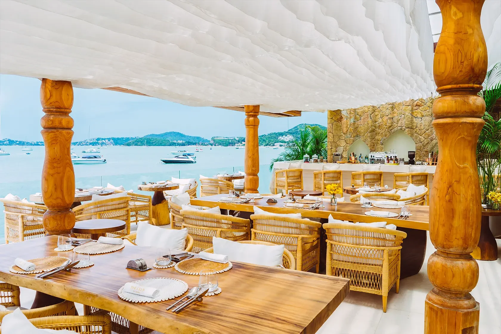 Upscale seaside restaurant with wooden tables, wicker chairs with white cushions, overlooking a bay with boats and green hills, stone and wood decor, and a bar area in the background.