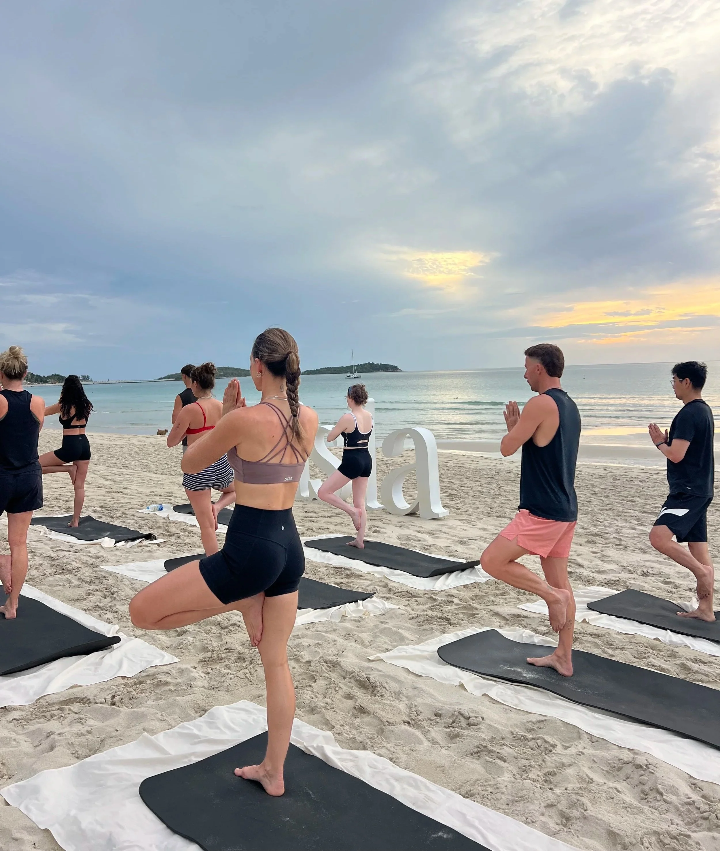People practicing yoga on black mats on a sandy beach with the ocean and an island in the background, during sunset or early evening.