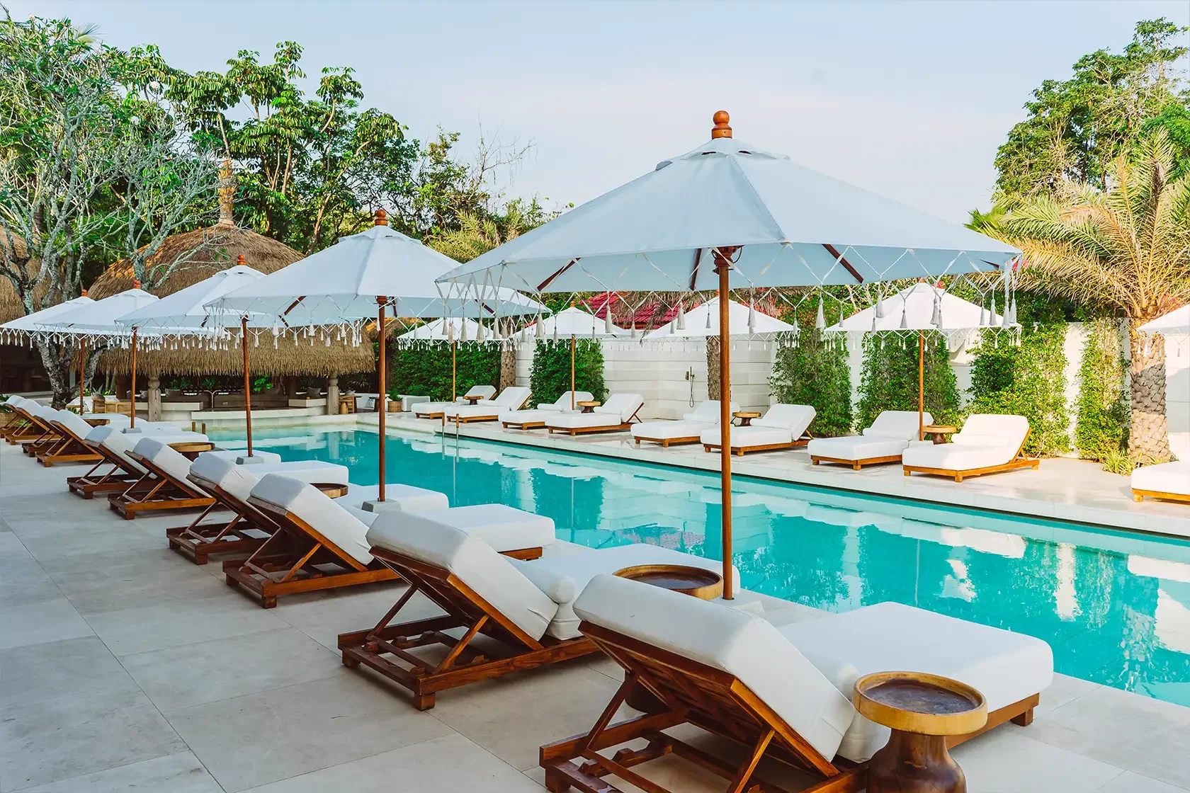 Luxury swimming pool area with white lounge chairs and umbrellas, surrounded by greenery and thatched-roof huts.
