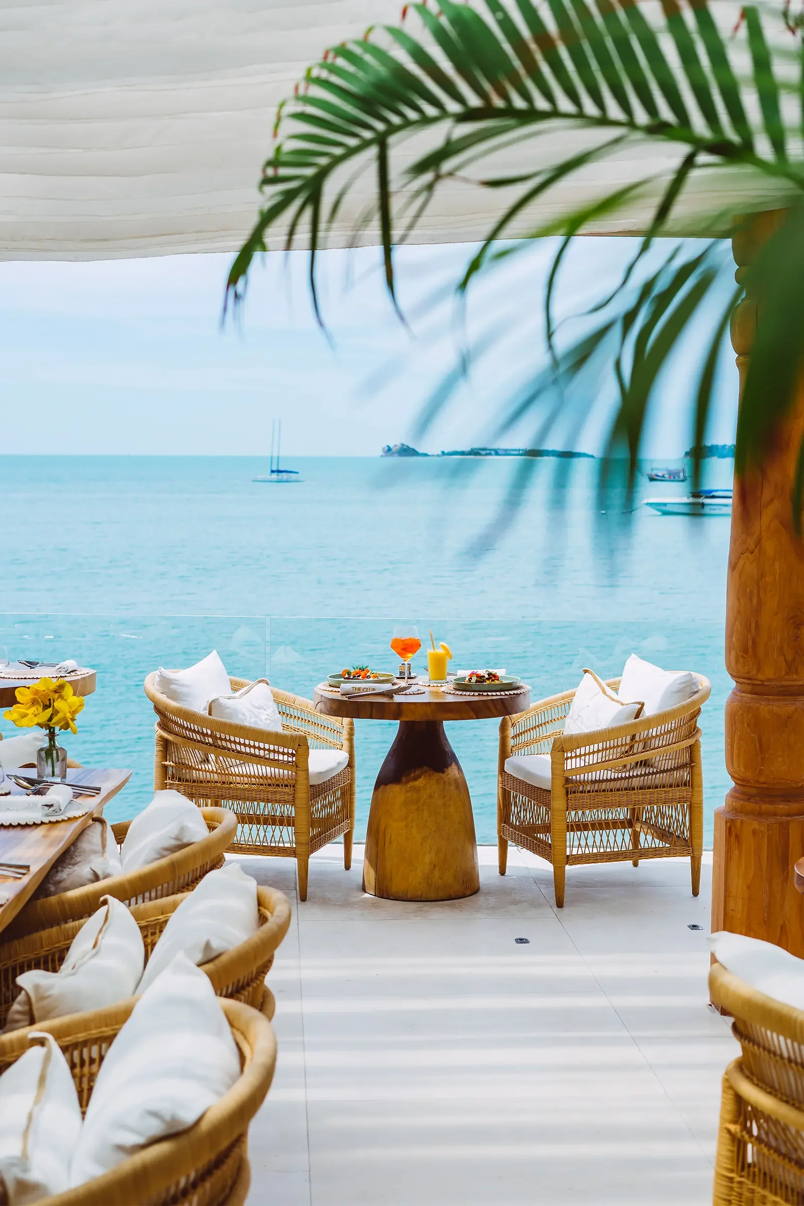 A seaside restaurant or café with wooden tables and seating, decorated with white pillows and yellow flowers, overlooking the ocean with sailboats and a distant shoreline.