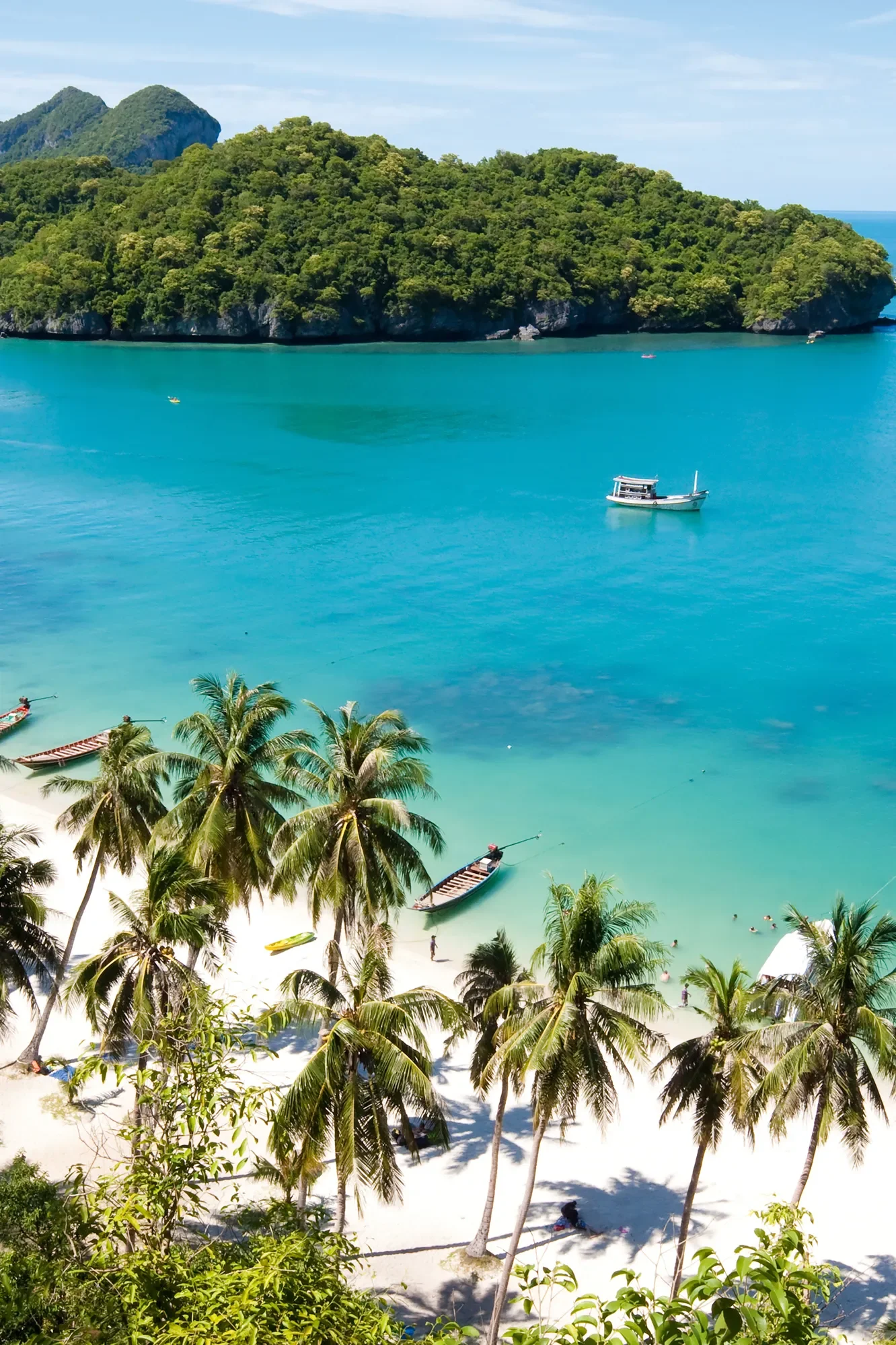 A tropical beach with white sand, turquoise water, and lush palm trees. Several boats are floating on the water, and there are hills covered in green vegetation in the background.