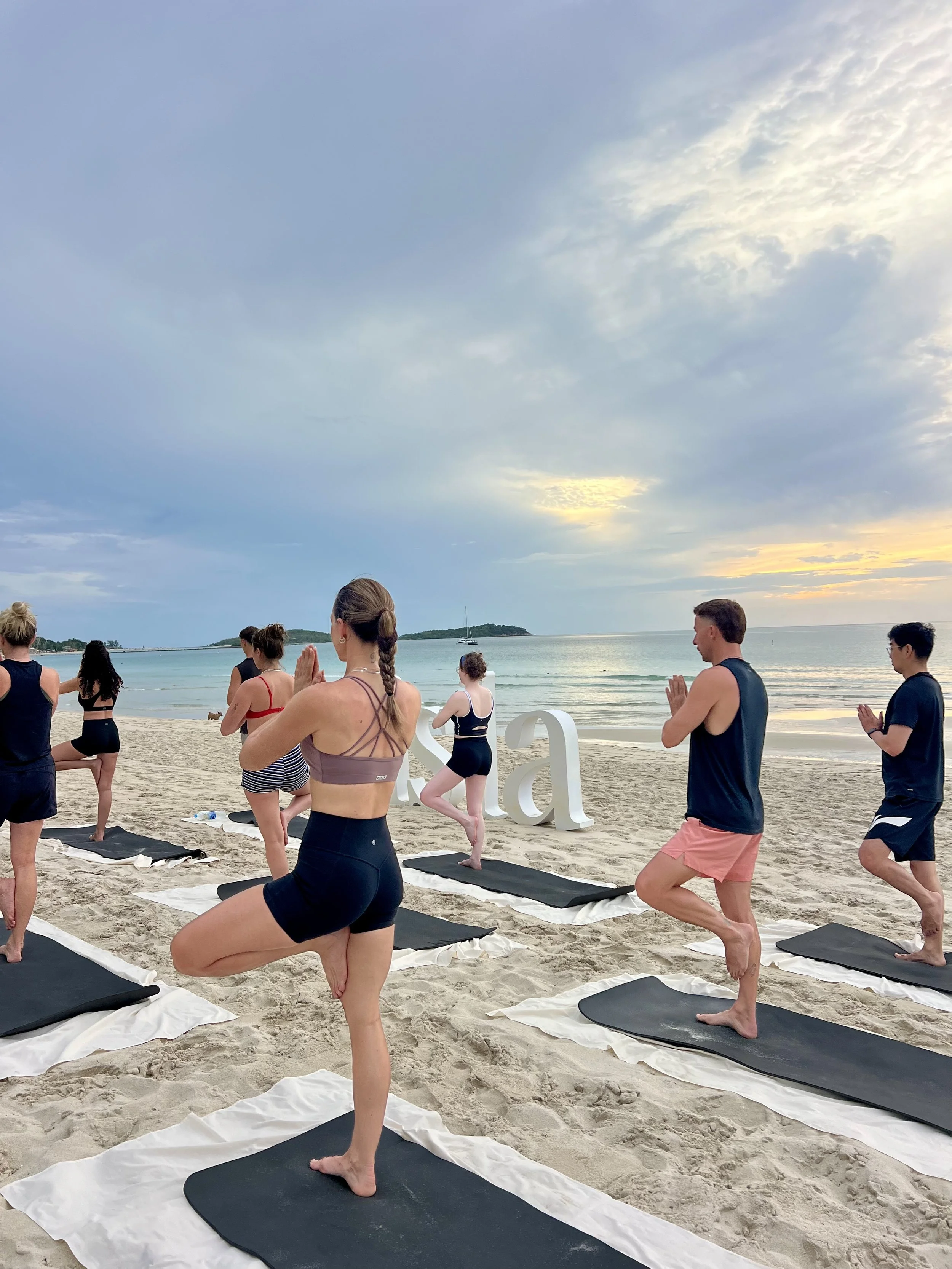 People practicing yoga on a sandy beach during sunset, with the ocean and small island in the background.