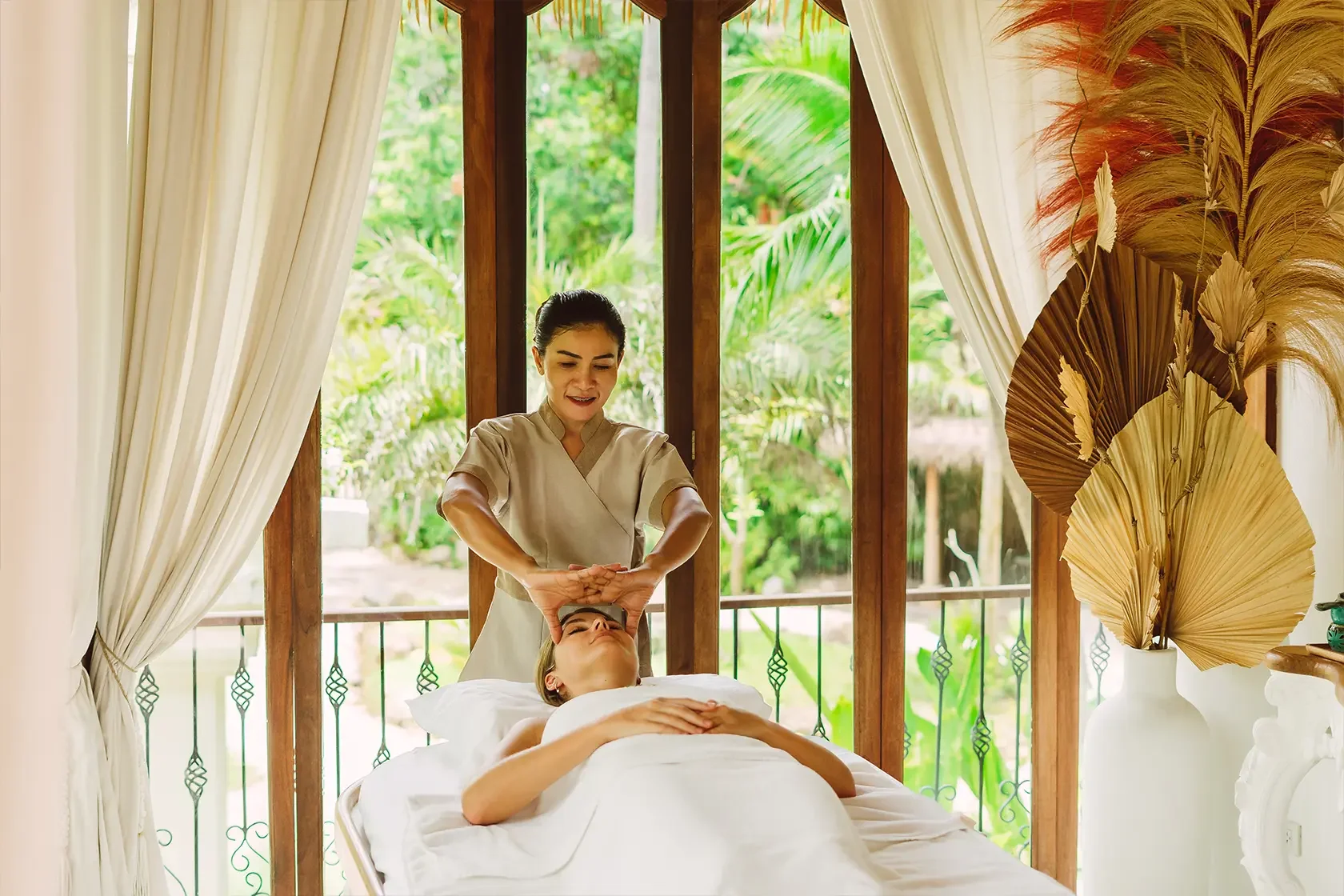 A woman receiving a facial treatment from a massage therapist in a serene, sunlit room decorated with large dried leaves in a vase and white curtains, with lush greenery visible outside.