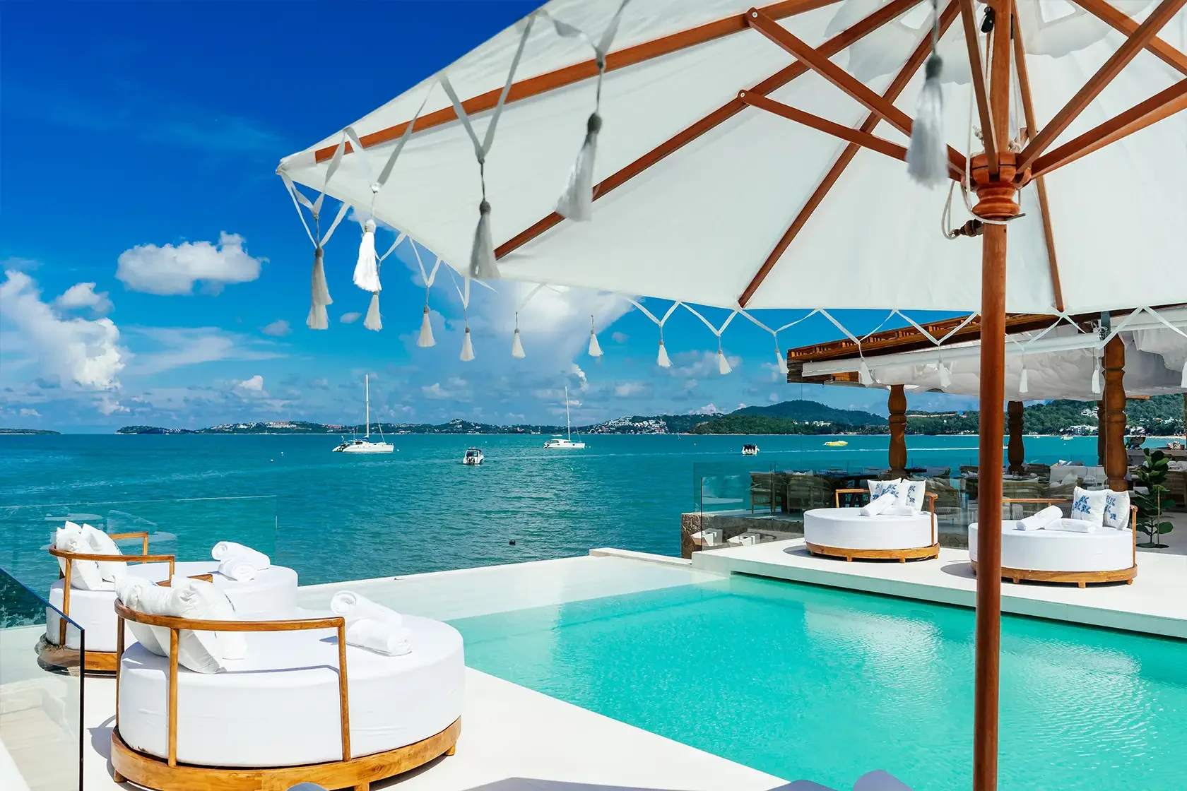 Luxury outdoor pool area overlooking the ocean with white cushioned lounge chairs, wooden umbrellas, and sailboats in the water under a clear blue sky.