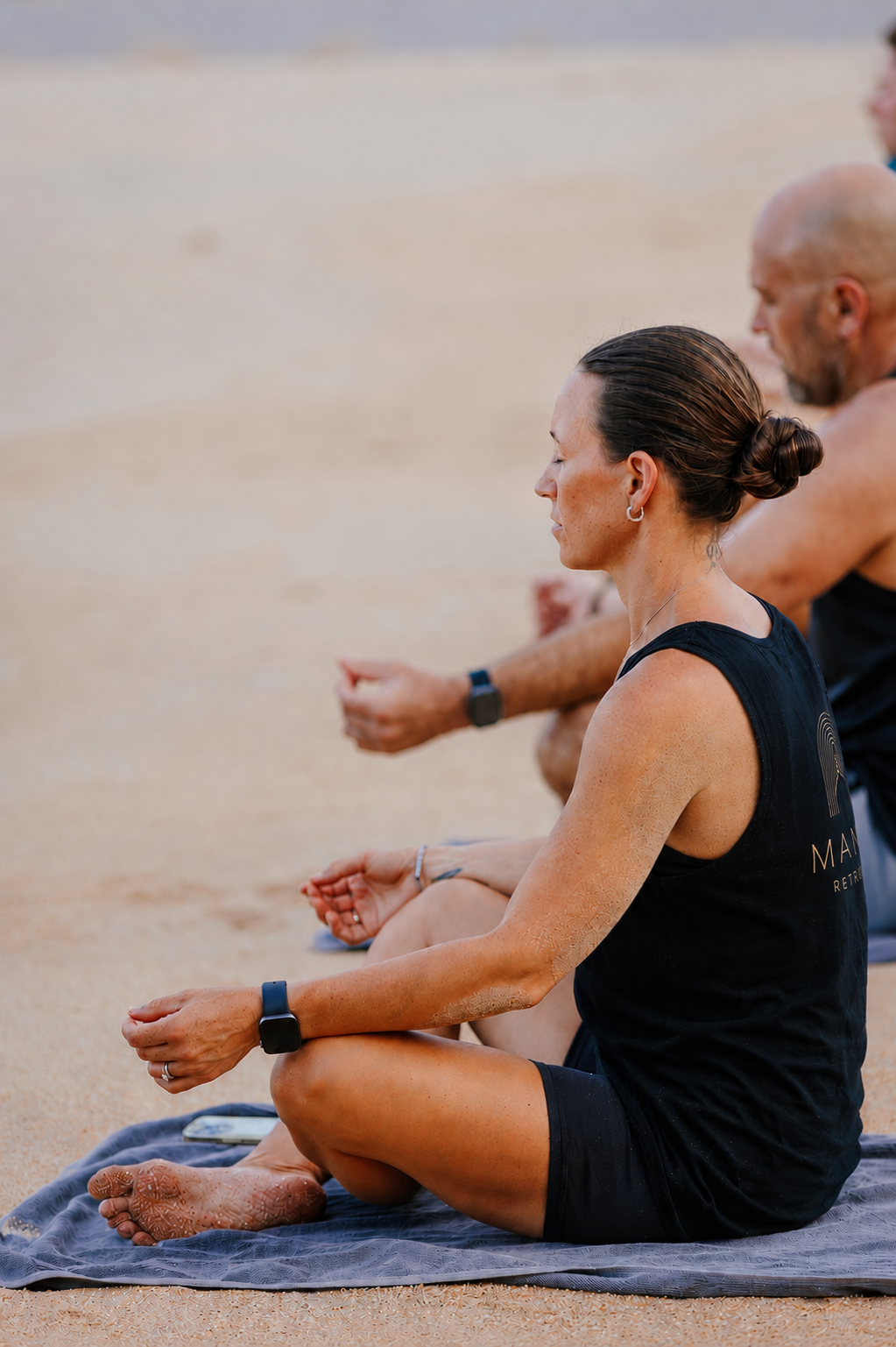 A group of people sitting cross-legged on towels on the sand, practicing meditation with eyes closed, in an outdoor setting.