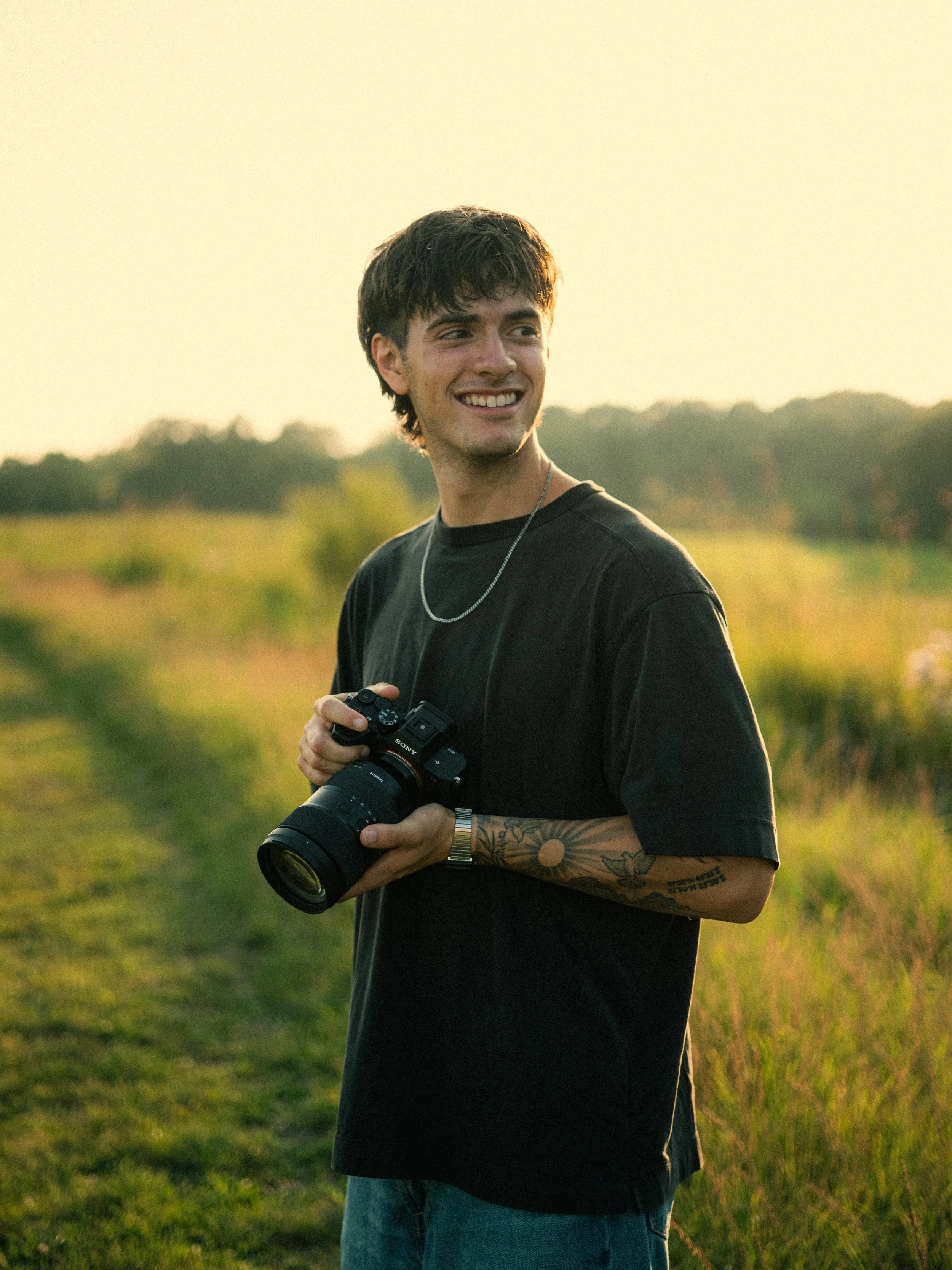 A young man with dark hair, tattoos on his left arm, smiling and holding a camera in a field during sunset.