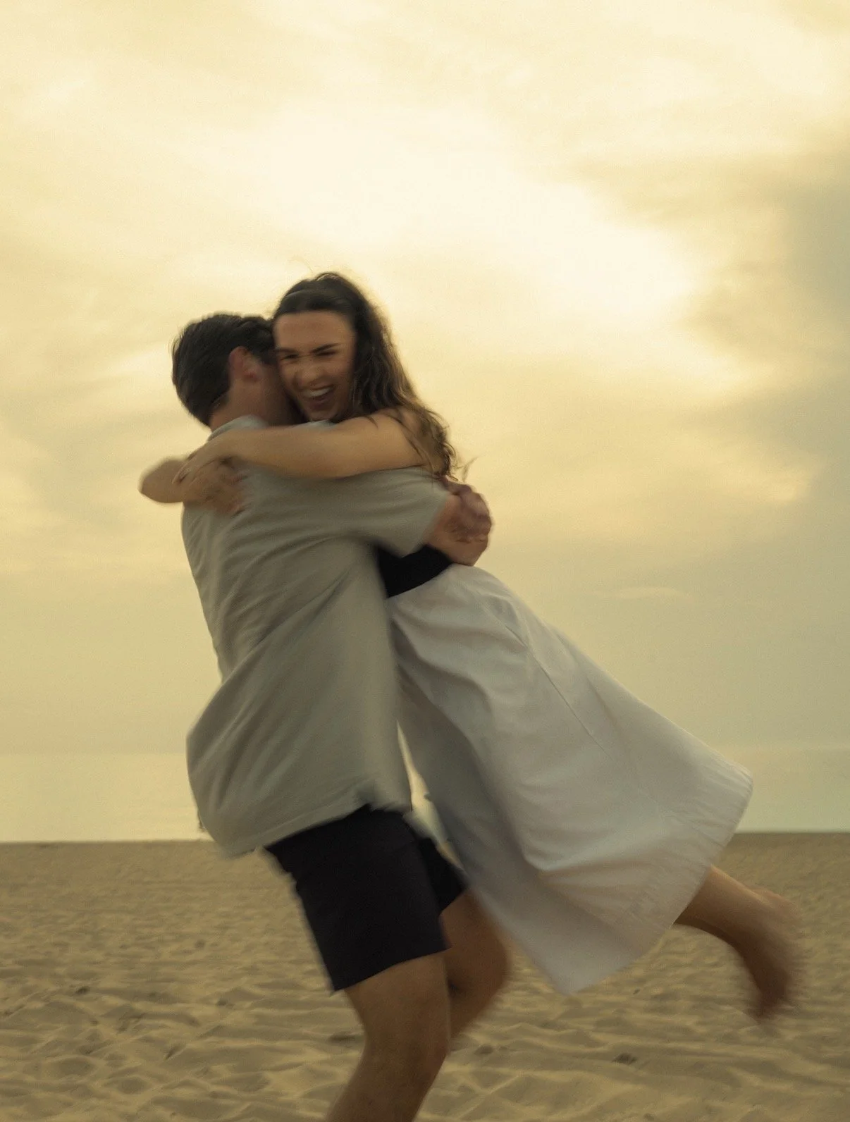 A man in shorts and a shirt lifting a woman in a dress on a beach during sunset.