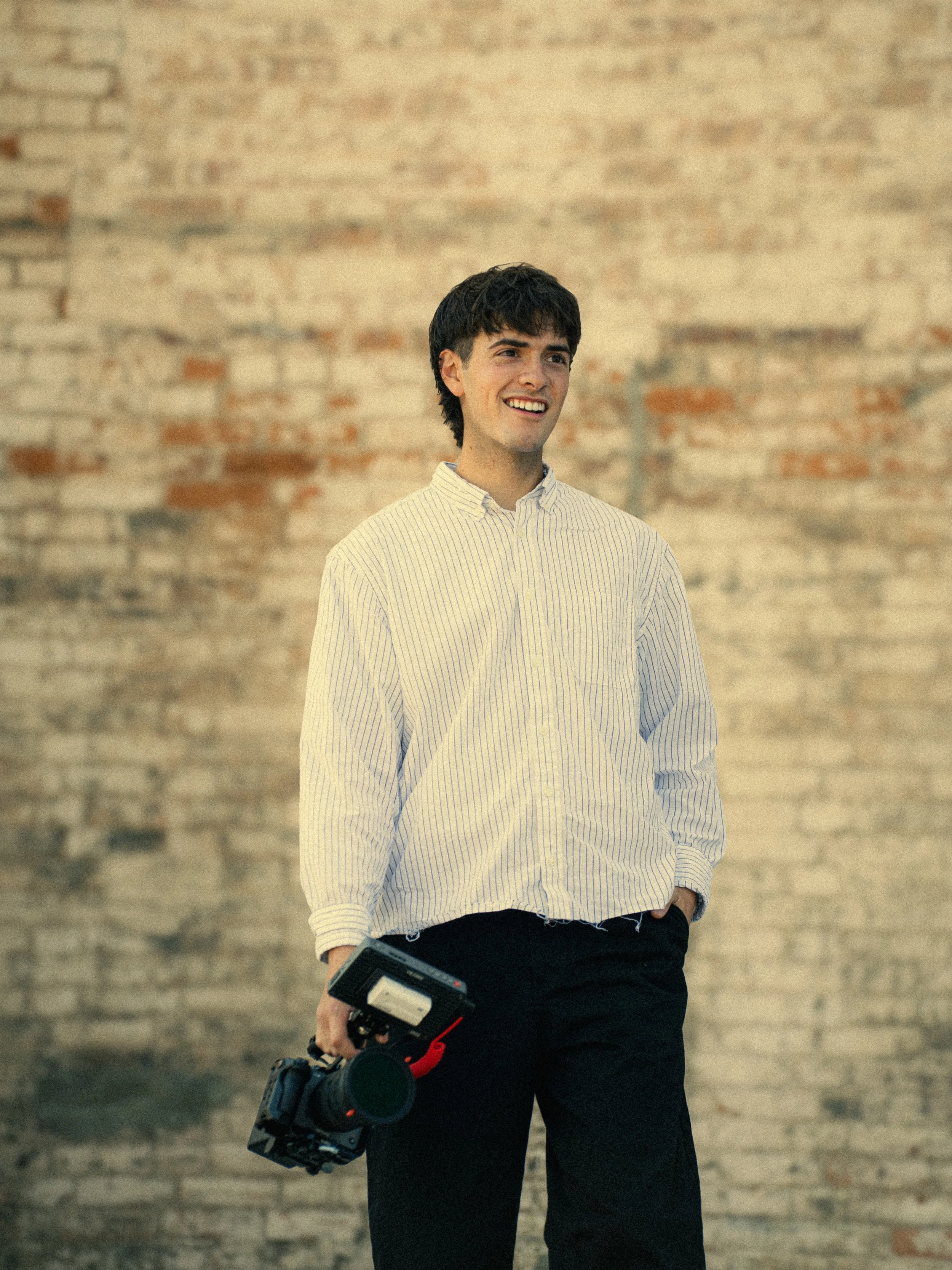 A young man with dark hair wearing a white and yellow striped button-up shirt, holding a black camera and a small electronic device, standing in front of a textured brick wall.