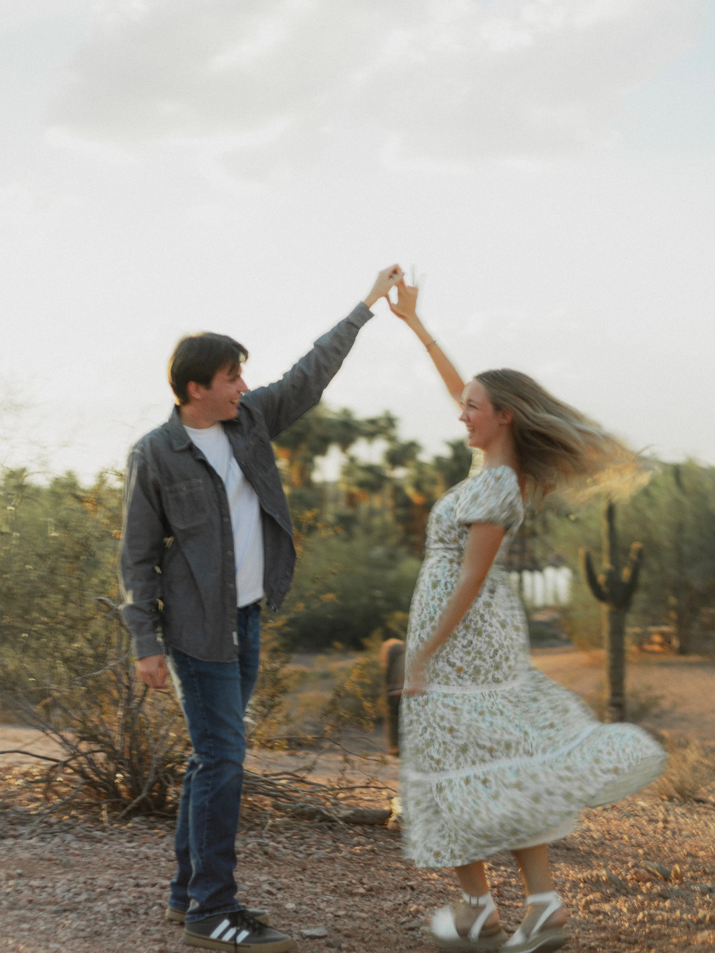 A young man and woman are dancing and holding hands outdoors during sunset. The woman is wearing a floral dress and white shoes, and the man is dressed in jeans, a white t-shirt, a gray jacket, and sneakers. They are smiling and appear to be enjoying
