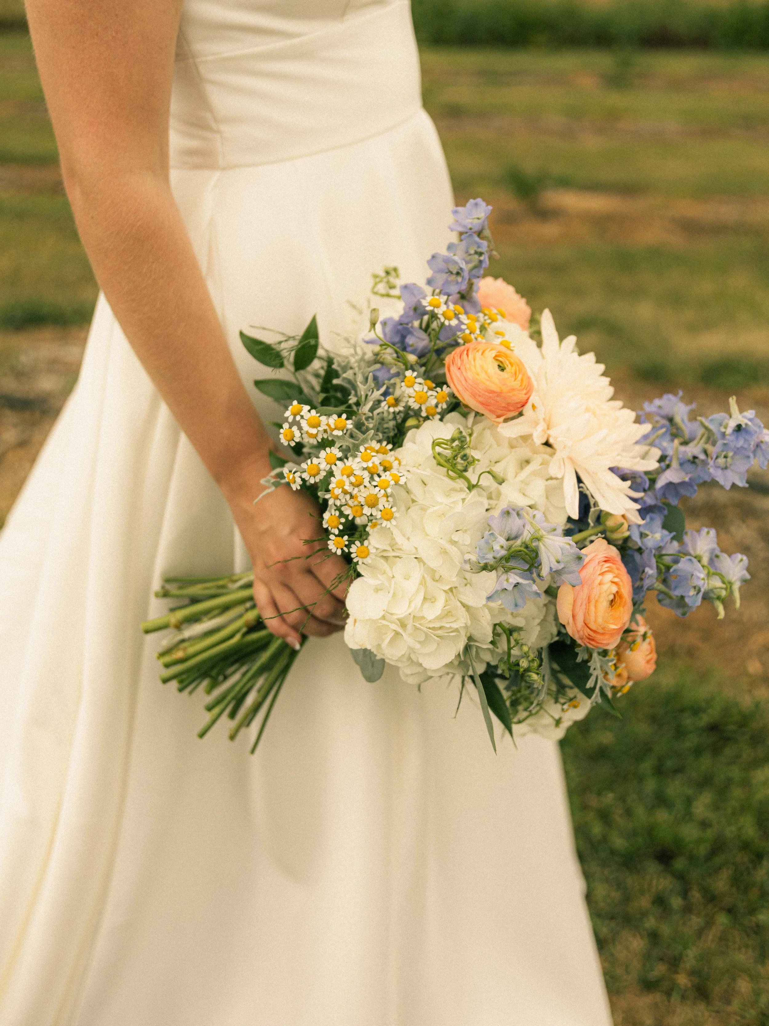 A person in a white dress holding a bouquet of mixed flowers including white hydrangeas, peach ranunculus, purple delphiniums, and small white daisies with yellow centers.