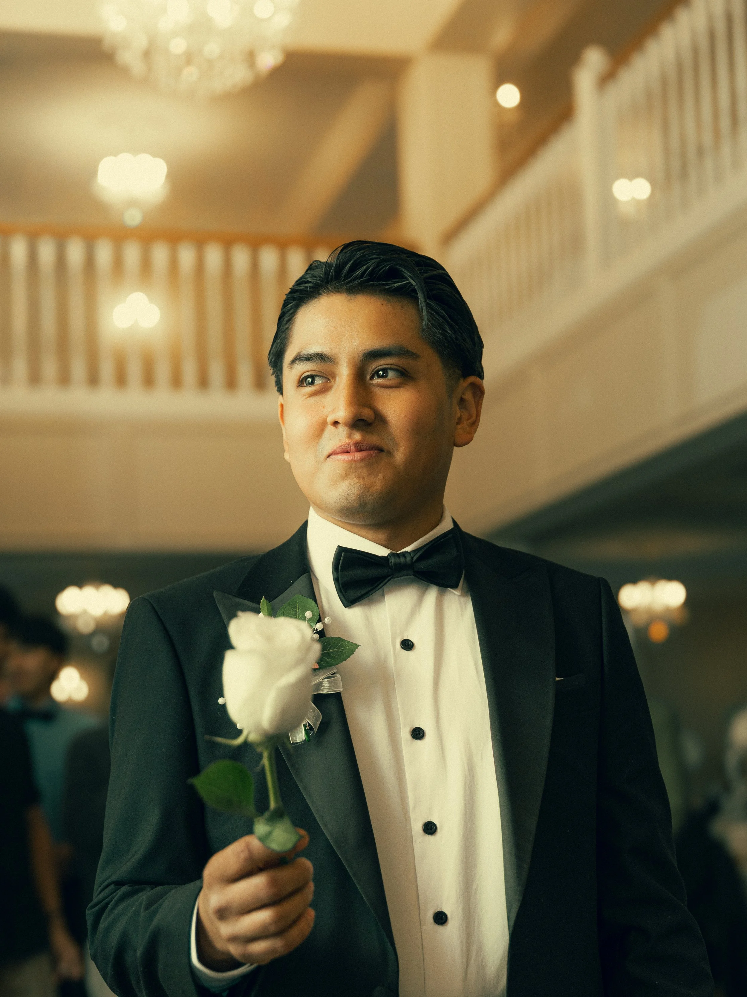 A man in a tuxedo holding a white rose at a formal event or wedding reception.