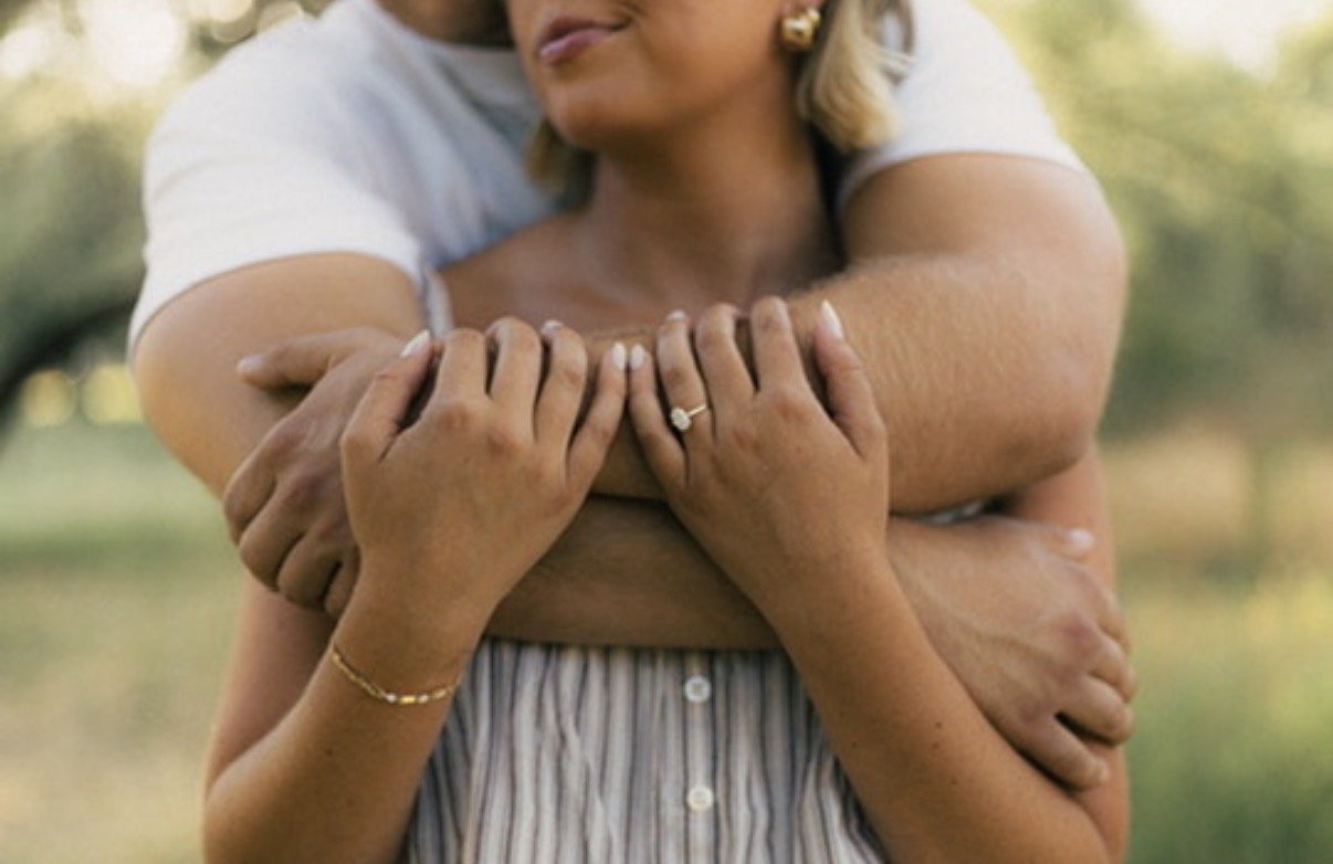 A woman wearing a white shirt being hugged from behind by a man, with her arms crossed in front of her chest, outdoors with blurred trees and grass in the background.