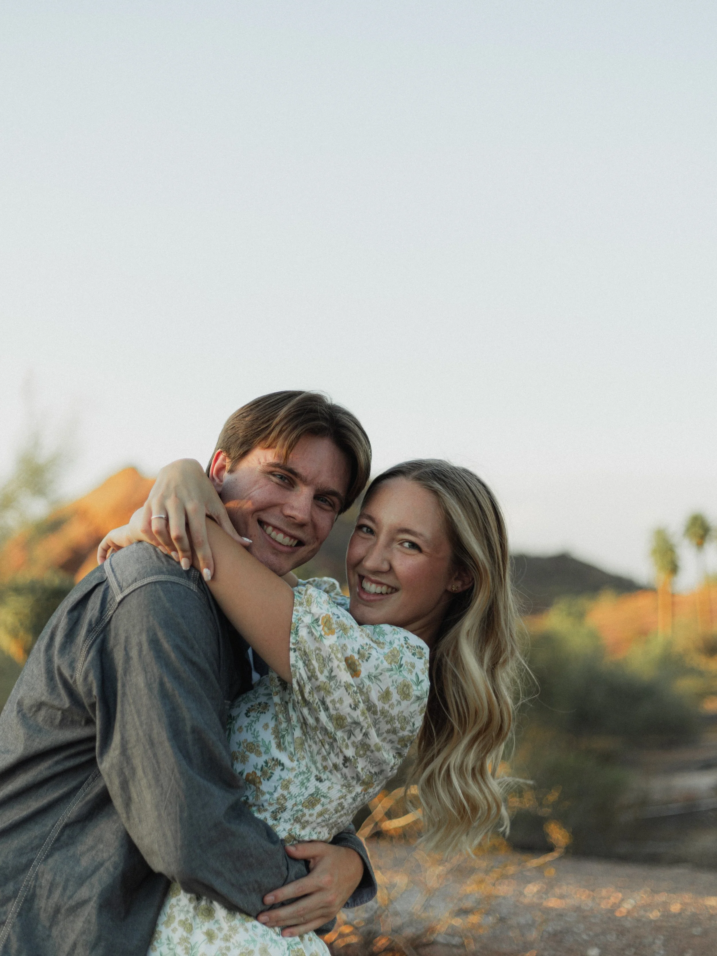 A smiling man and woman hugging outdoors during sunset with autumn trees and mountains in the background.