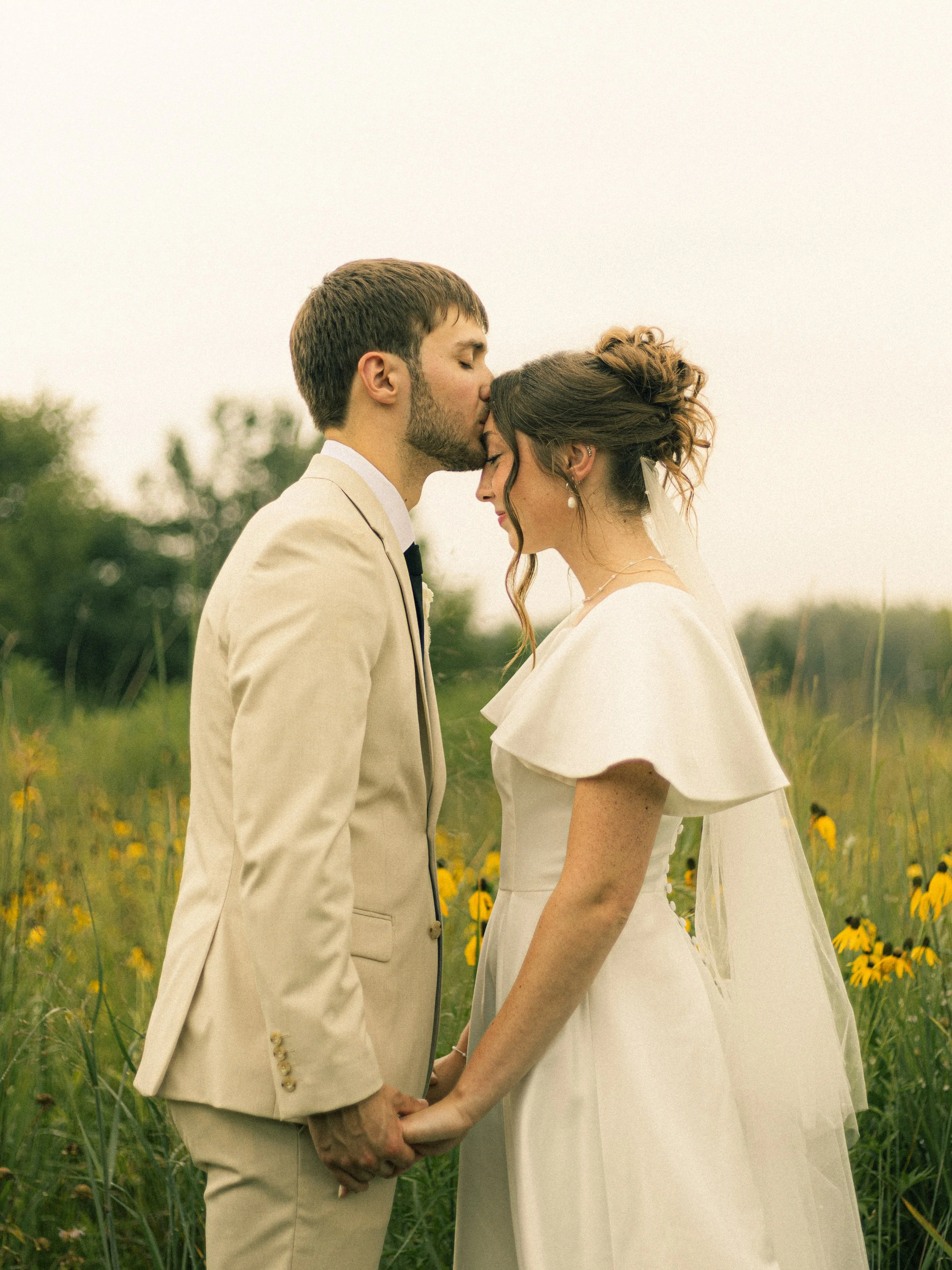 A bride and groom stand close together in a field of yellow flowers, holding hands and sharing a tender moment with their foreheads touching and eyes closed.